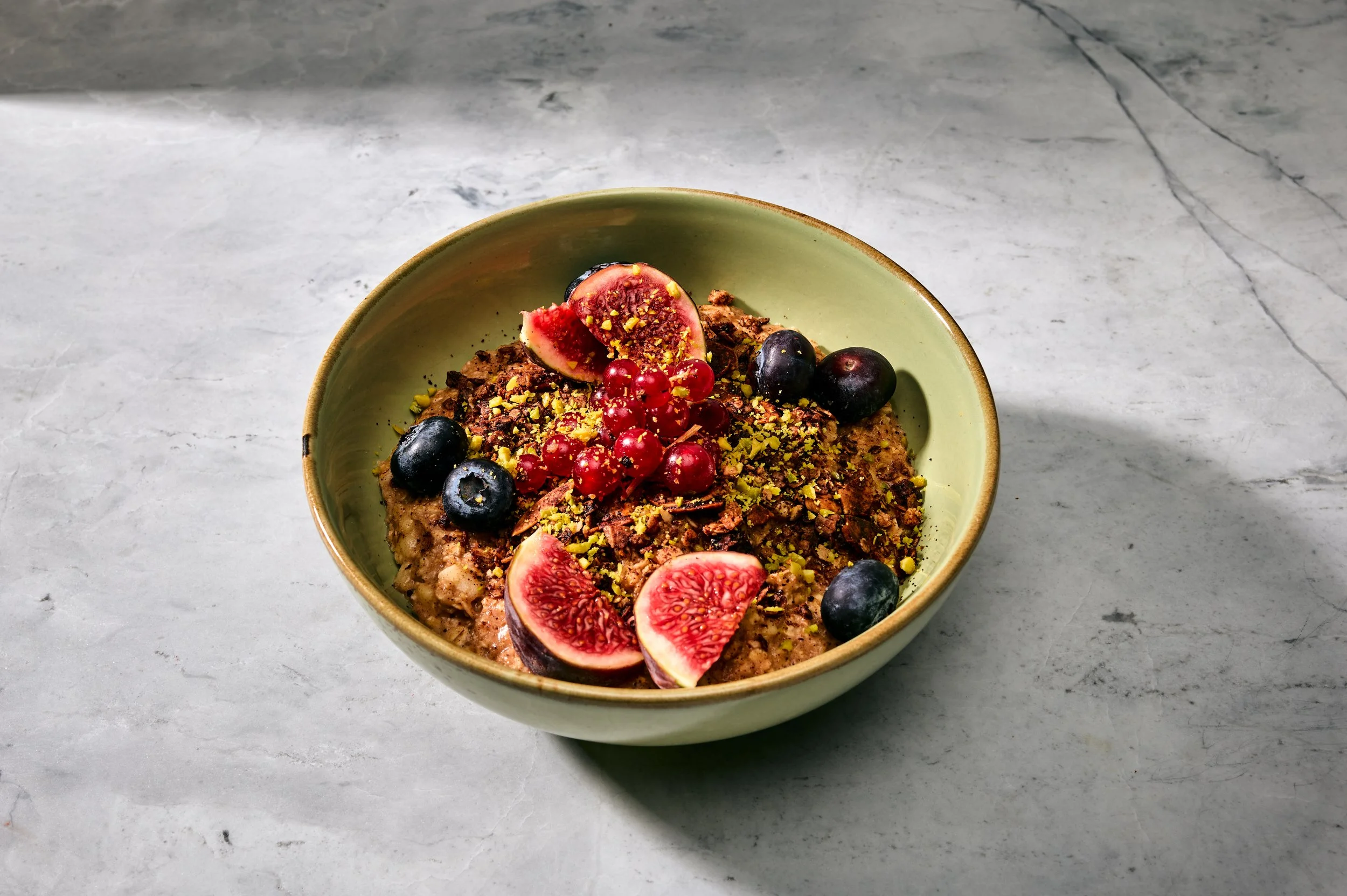 Schüssel mit Müsli, belegt mit Blaubeeren, geschnittenen Feigen und Pistazien, auf einer grauen Mamorplatte.
