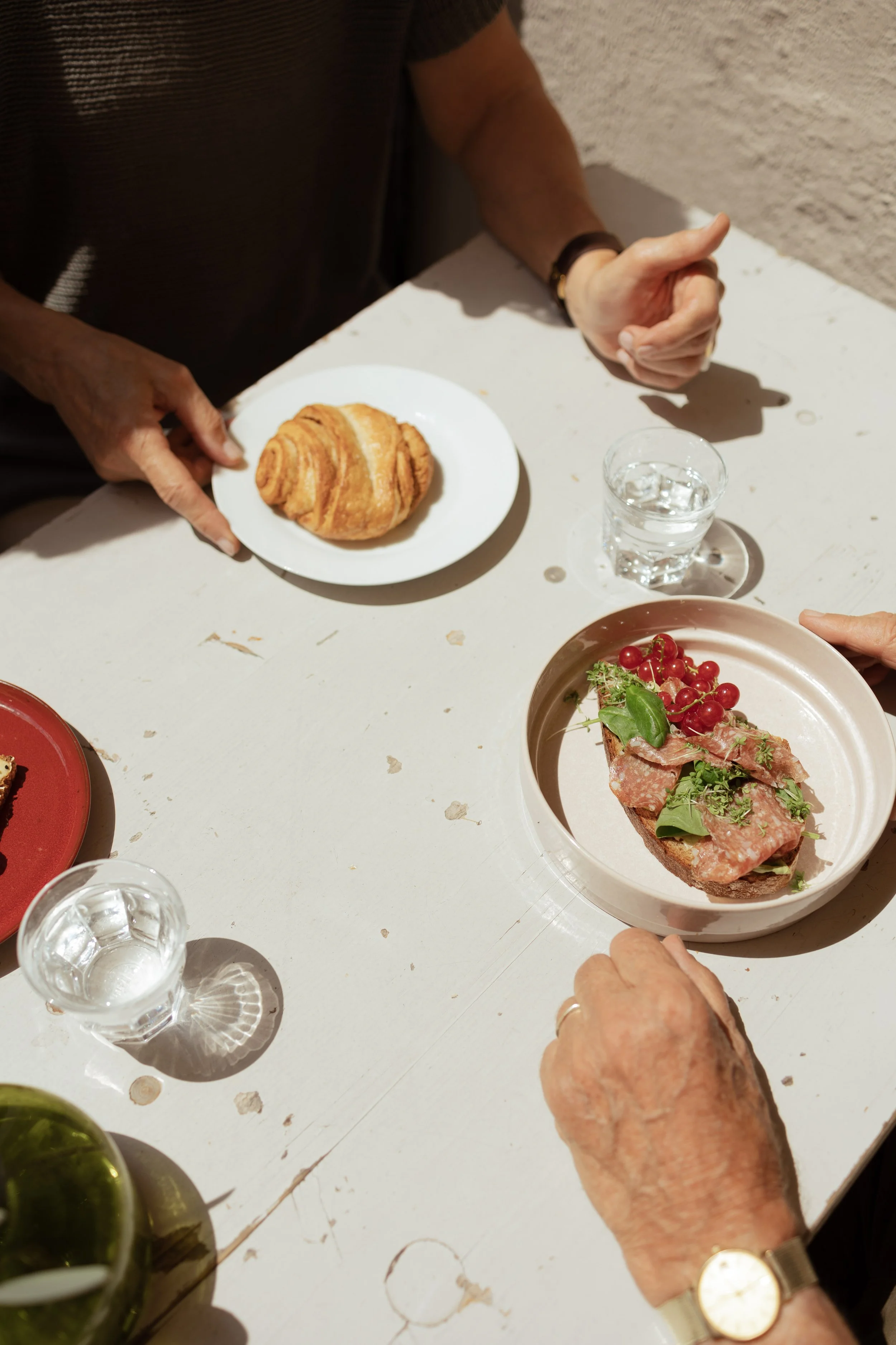 Ältere Person hält eine Scheibe Sauerteigbrot mit Aufschnitt und Kirschtomaten, während eine andere Personen ein Franzbrötchen bestellt hat.