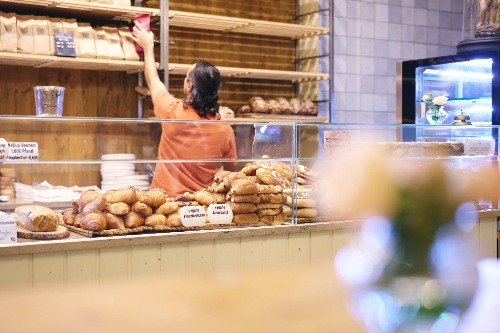 Bäcker verkauft vegane Croissants und französische Brötchen in einer Bäckerei. Im Hintergrund stehen Brote auf Regalen, und eine Verkäuferin richtet die Waren zurecht.