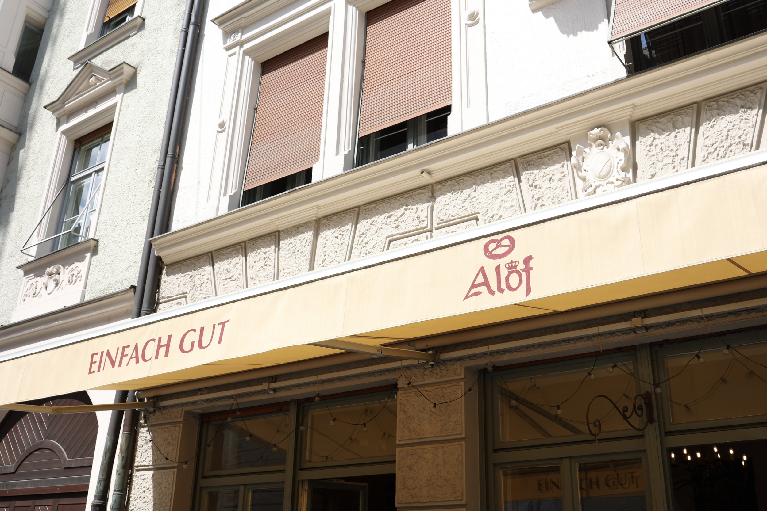 Ansicht eines Gebäudes mit barocken Fassadendetails, schließbare Fensterläden und einer Ladenfront mit Sonnenschutzmarkise, die das Wort 'Alof' zeigt.