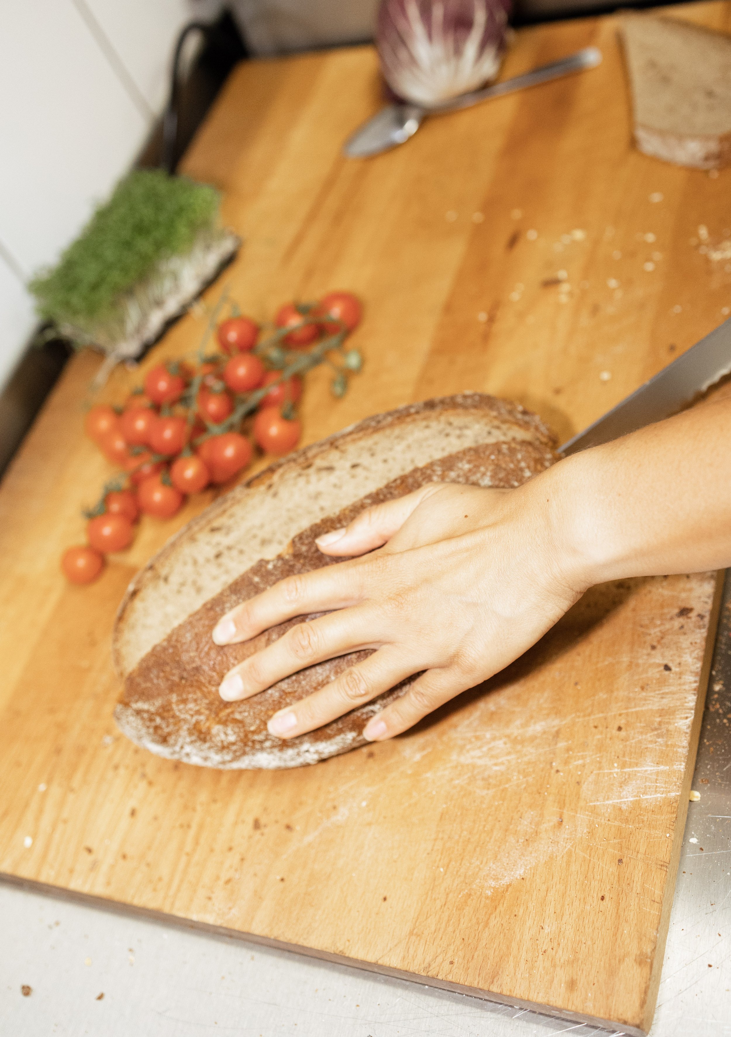 Eine Person schneidet ein Brot auf einem Holzbrett, im Hintergrund sind Tomaten auf der Ranke, ein Messer, ein Salat und ein Brotkanten zu sehen.