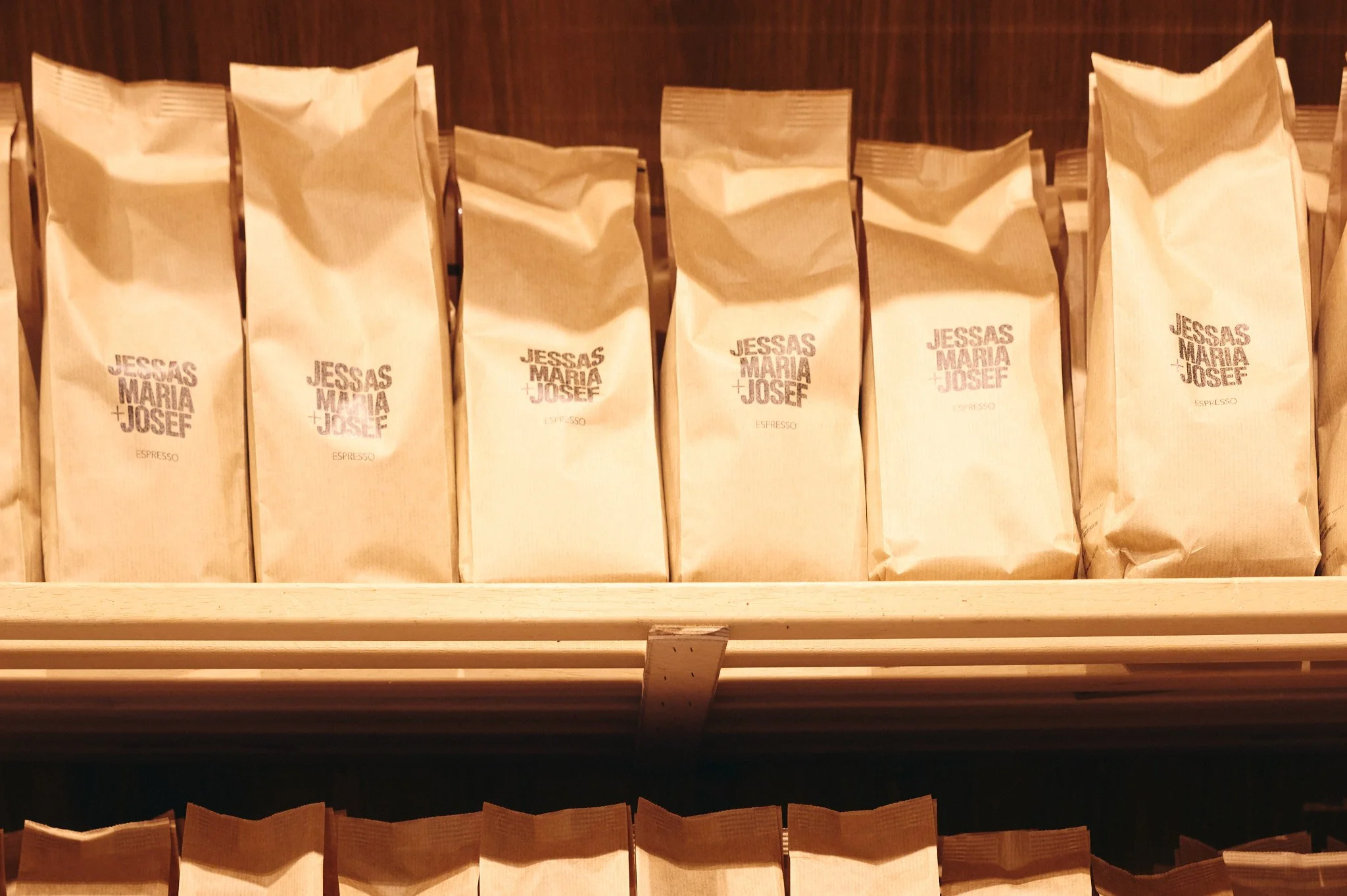 Mehrere braune Papiertüten mit Kaffeebohnen, einige mit Beschriftungen wie "Jessas Maria + Josef" und "Espresso", stehen in einem Regal.
