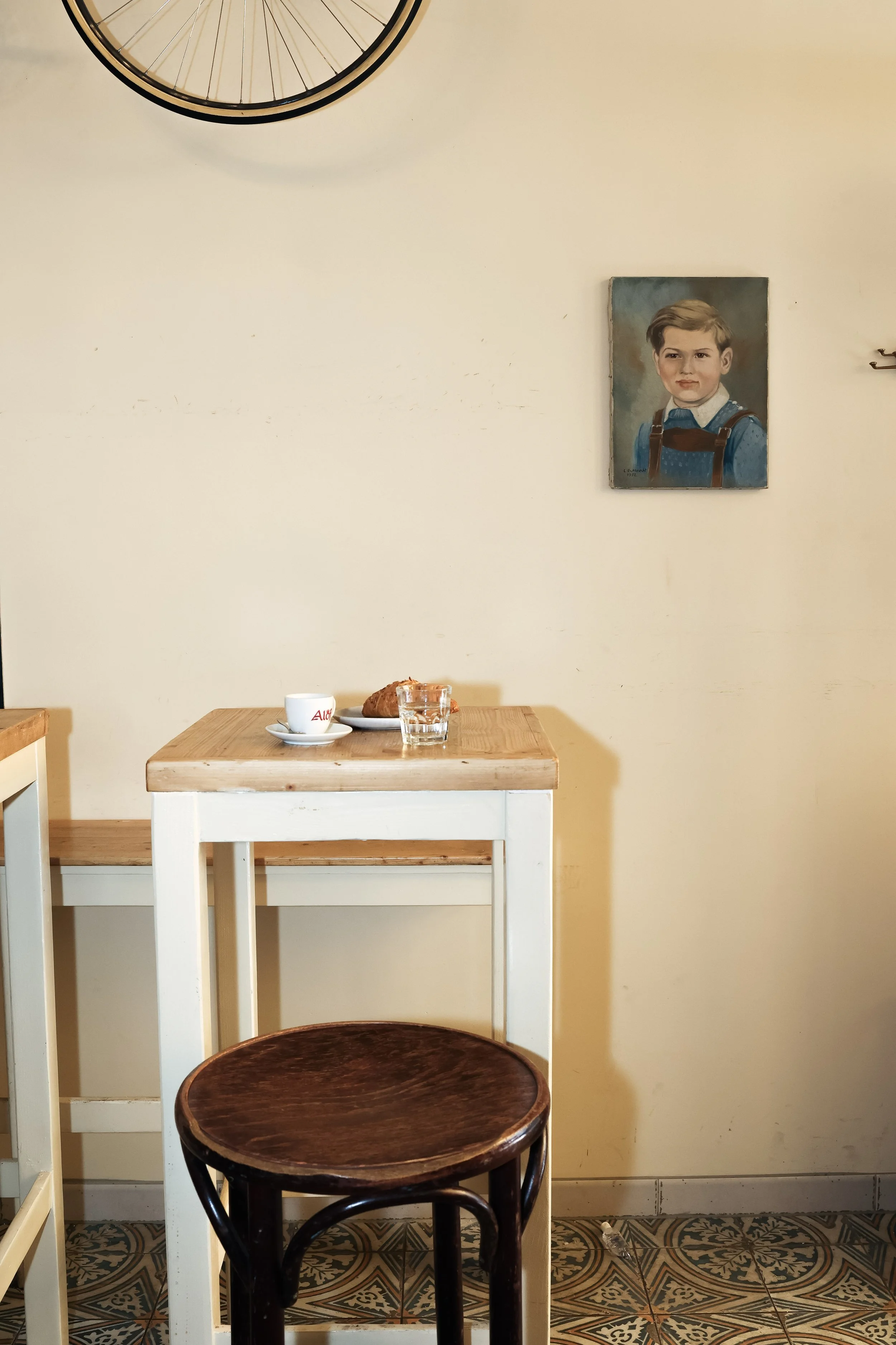 Ein heller Raum mit einem weißen Tisch, auf dem sich eine weiße Kaffeetasse, ein Glas Wasser und ein Stück Gebäck befinden. An der Wand hängt ein gemaltes Porträt eines Jungen. Im oberen linken Rand ist ein Fahrradreifen zu sehen. Der Boden besteht aus dekorativen Fliesen.