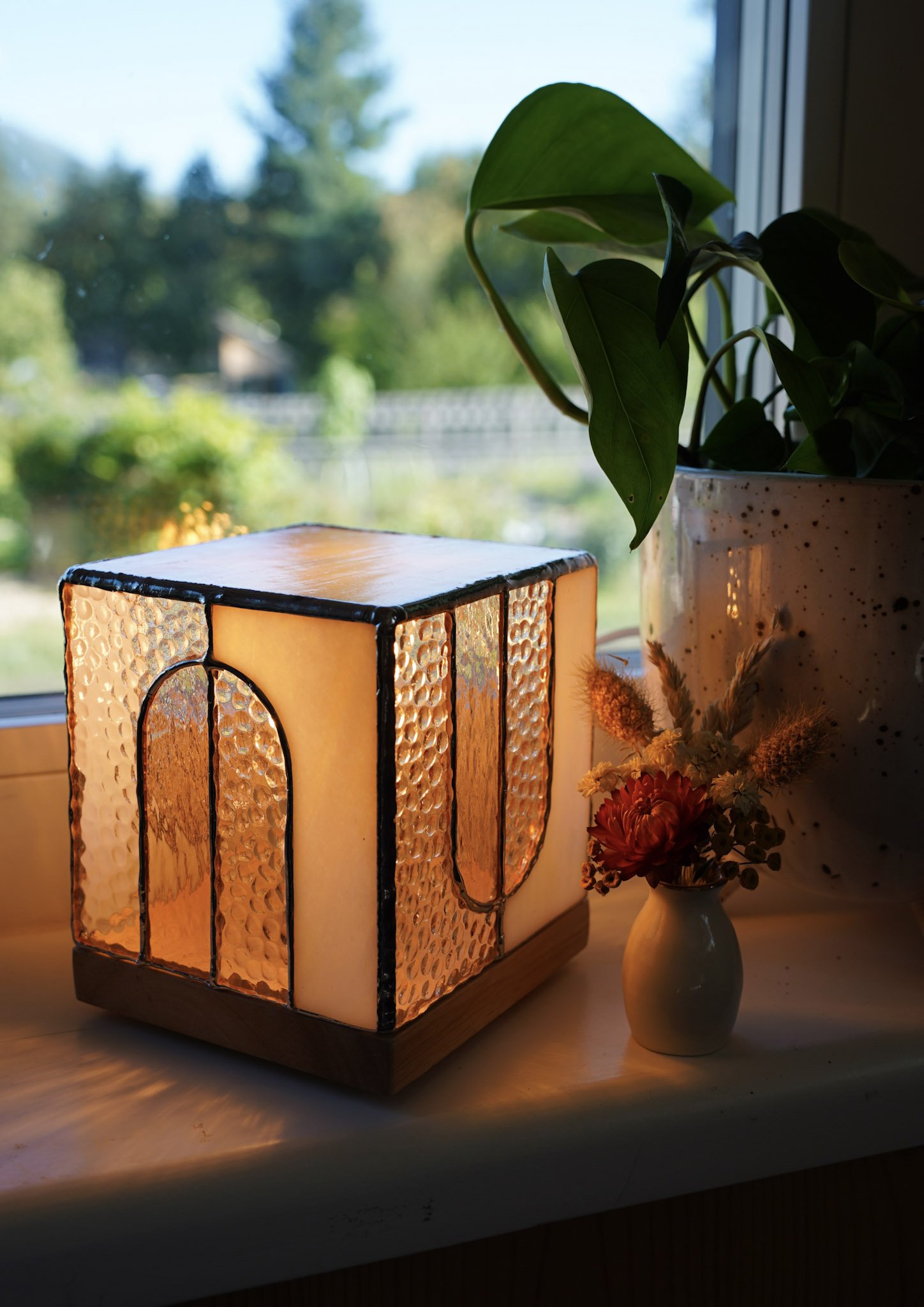 A lit stained glass lamp on a windowsill with a green potted plant and a small bouquet of flowers in a vase, with a blurred outdoor view of trees and a fence.