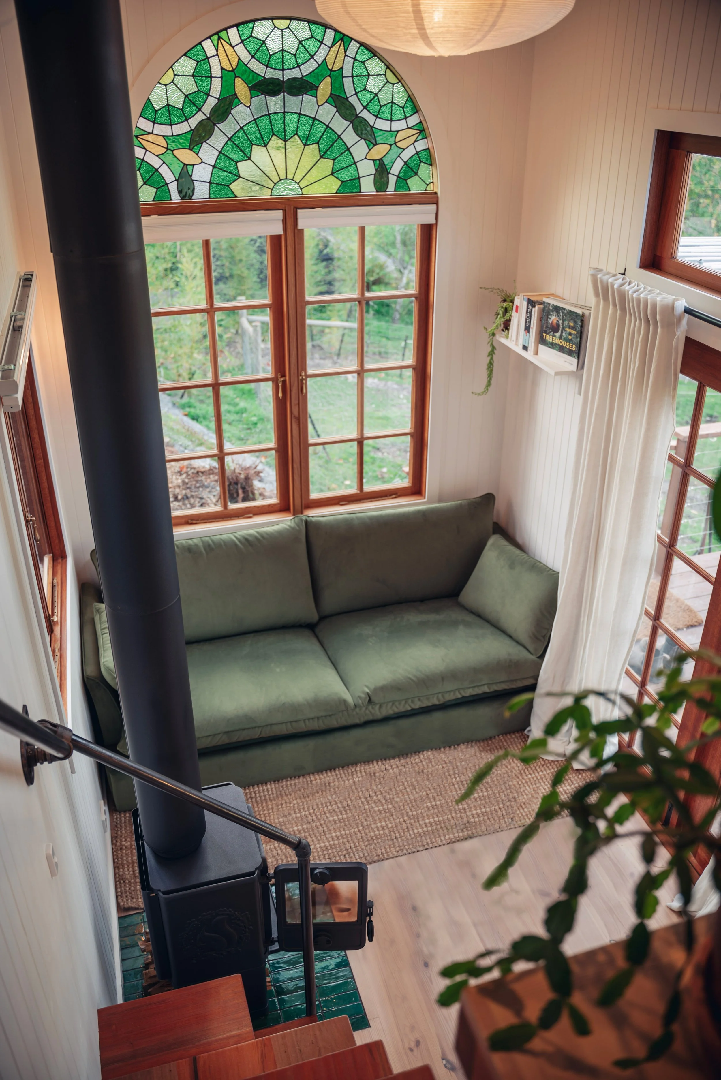 View of a living room with a sofa in front of large windows and a stained glass semicircular window at the top featuring green and blue geometric patterns and leaf motifs.