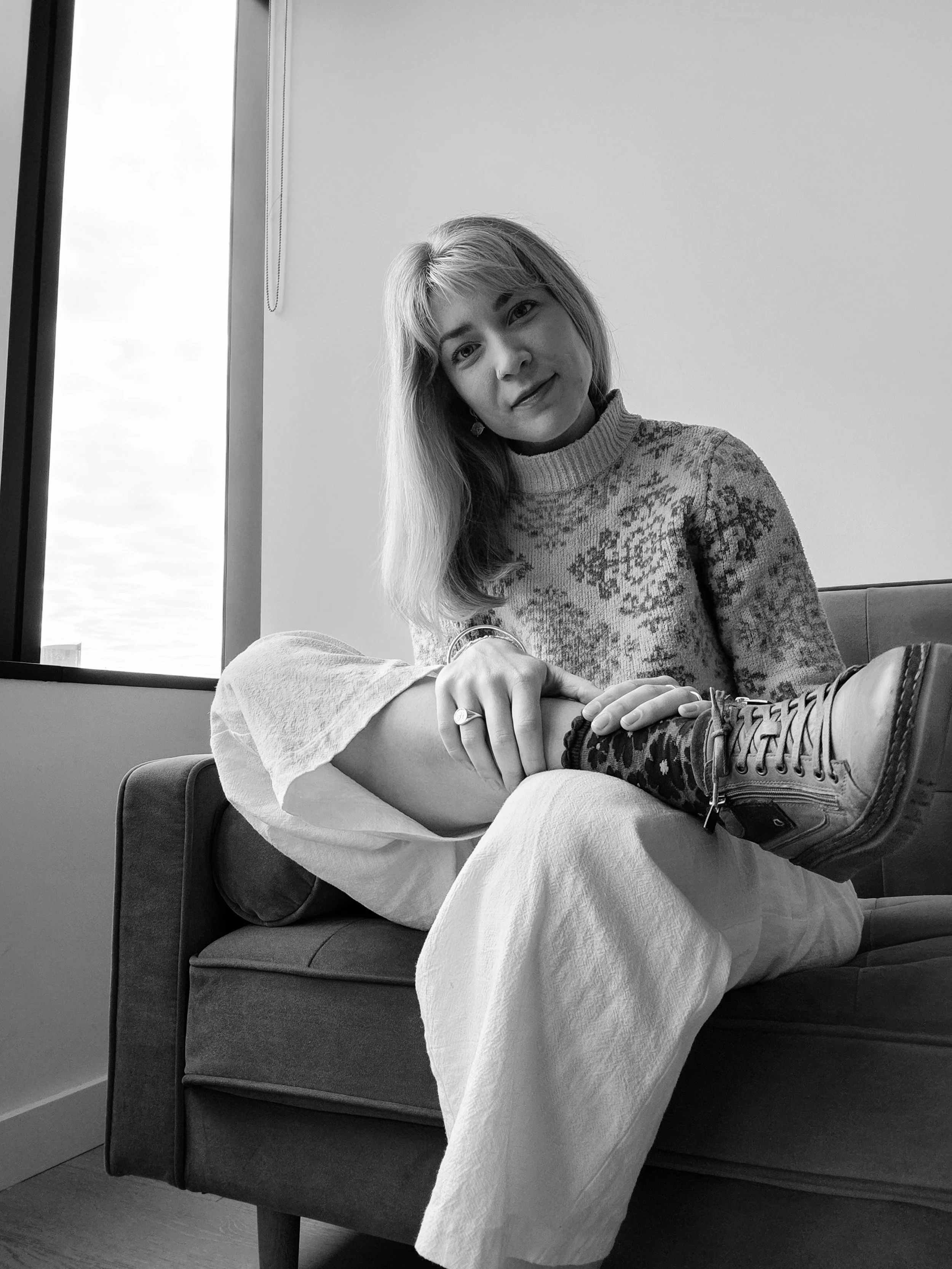 Black and white photo of a woman with long hair sitting on a couch, smiling at the camera. She is wearing a patterned sweater, a ring, and a bracelet. One of her shoes is visible on her foot. There is a window behind her showing the sky, and a chain hanging from the ceiling.