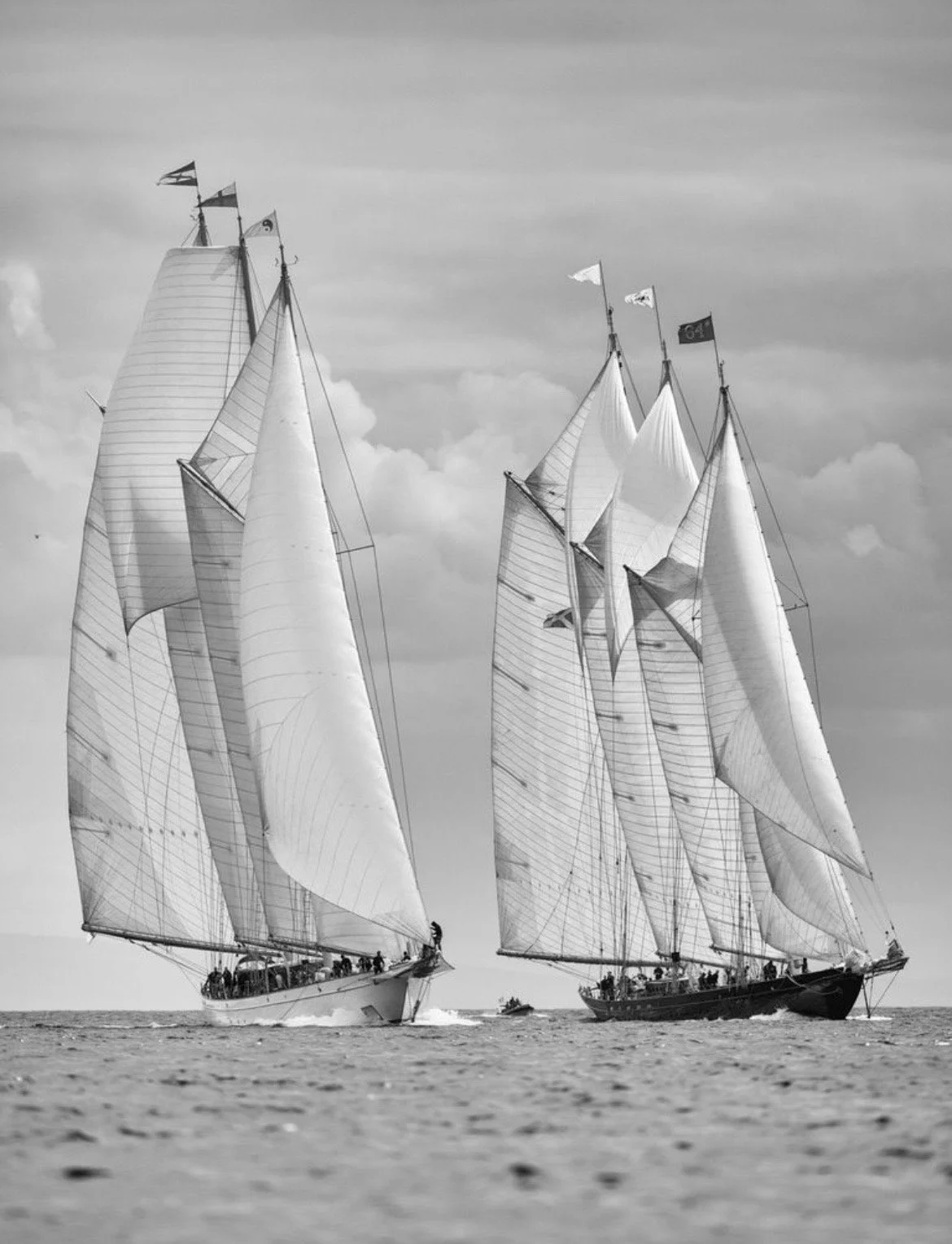 Two large sailboats with their sails fully raised, racing on the water during the day, with a cloudy sky in the background.