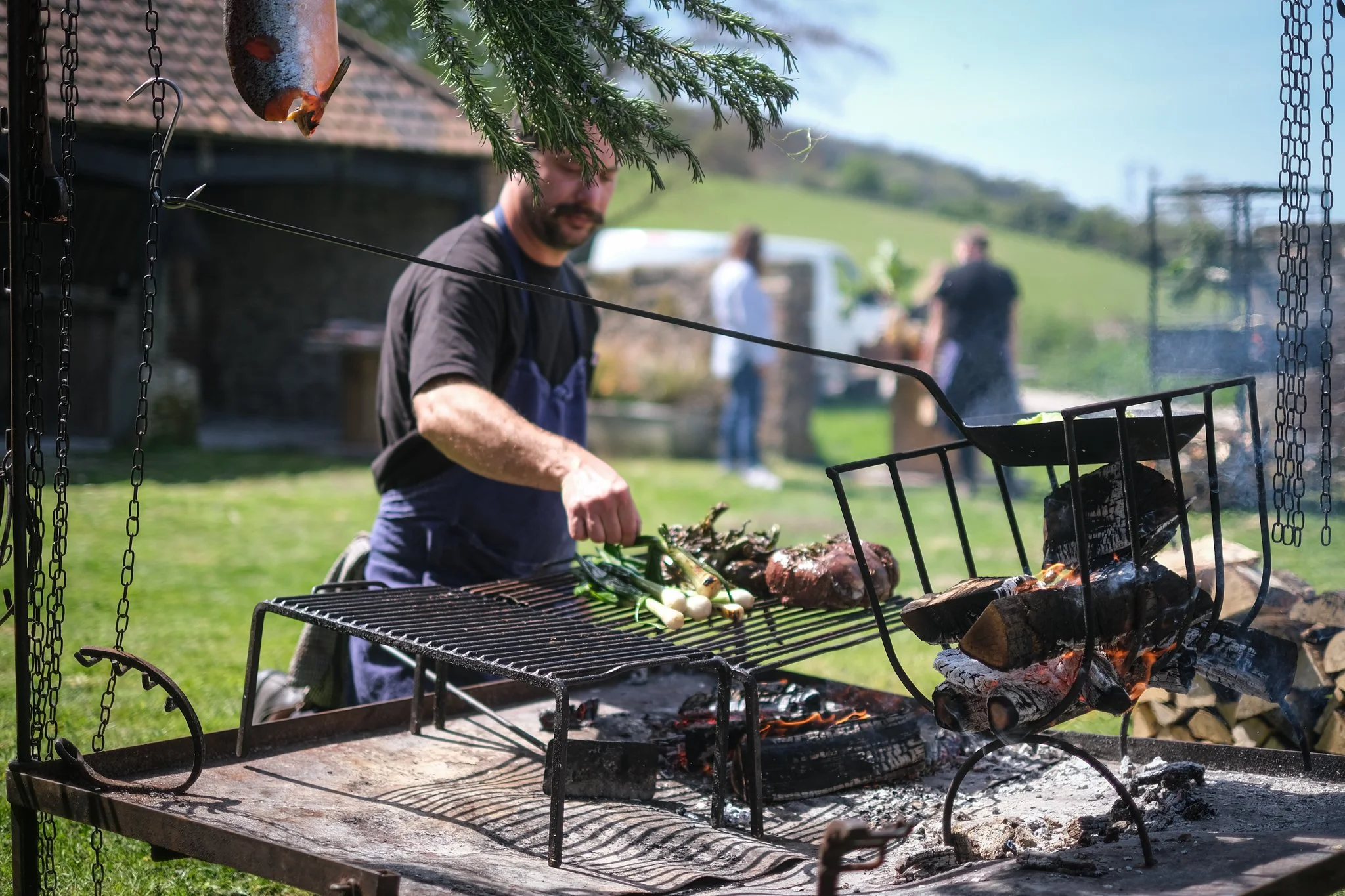 Man grilling vegetables and meat over an open fire outdoors, with people in the background and green hills in the distance.