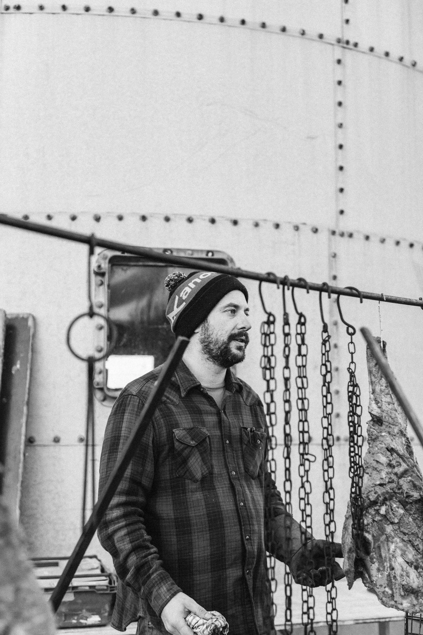 A man with a beard wearing a beanie and plaid shirt is handling a large piece of meat or animal carcass in a butchery or meat processing facility, with hooks and chains hanging in the background.