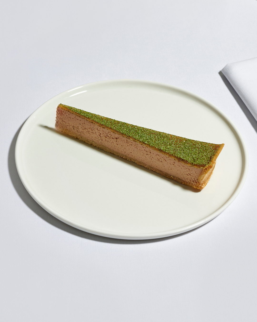 A slice of pink mousse cake with a green topping on a white oval plate, on a white table with a white napkin.