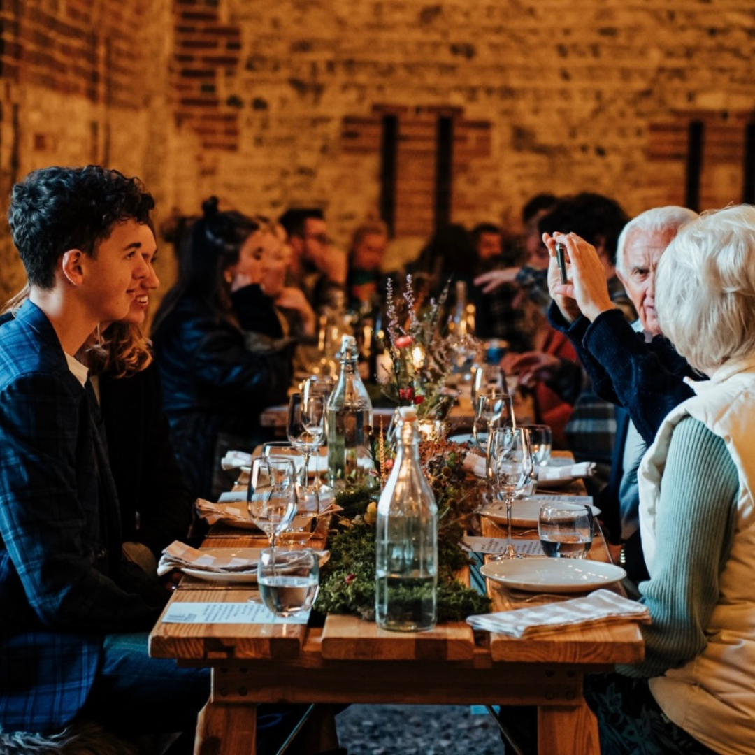 People sitting at a long dining table decorated with centerpieces, enjoying a meal in a rustic restaurant with exposed brick walls.