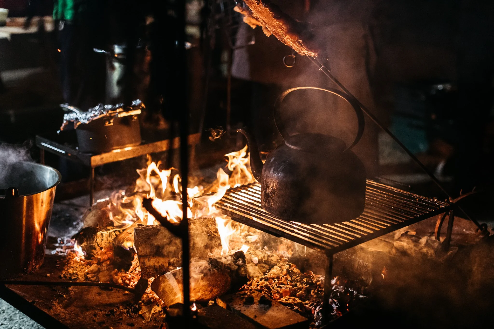 Cooking set over an open flame with a large pot on a grill, surrounded by burning wood and glowing embers at night.