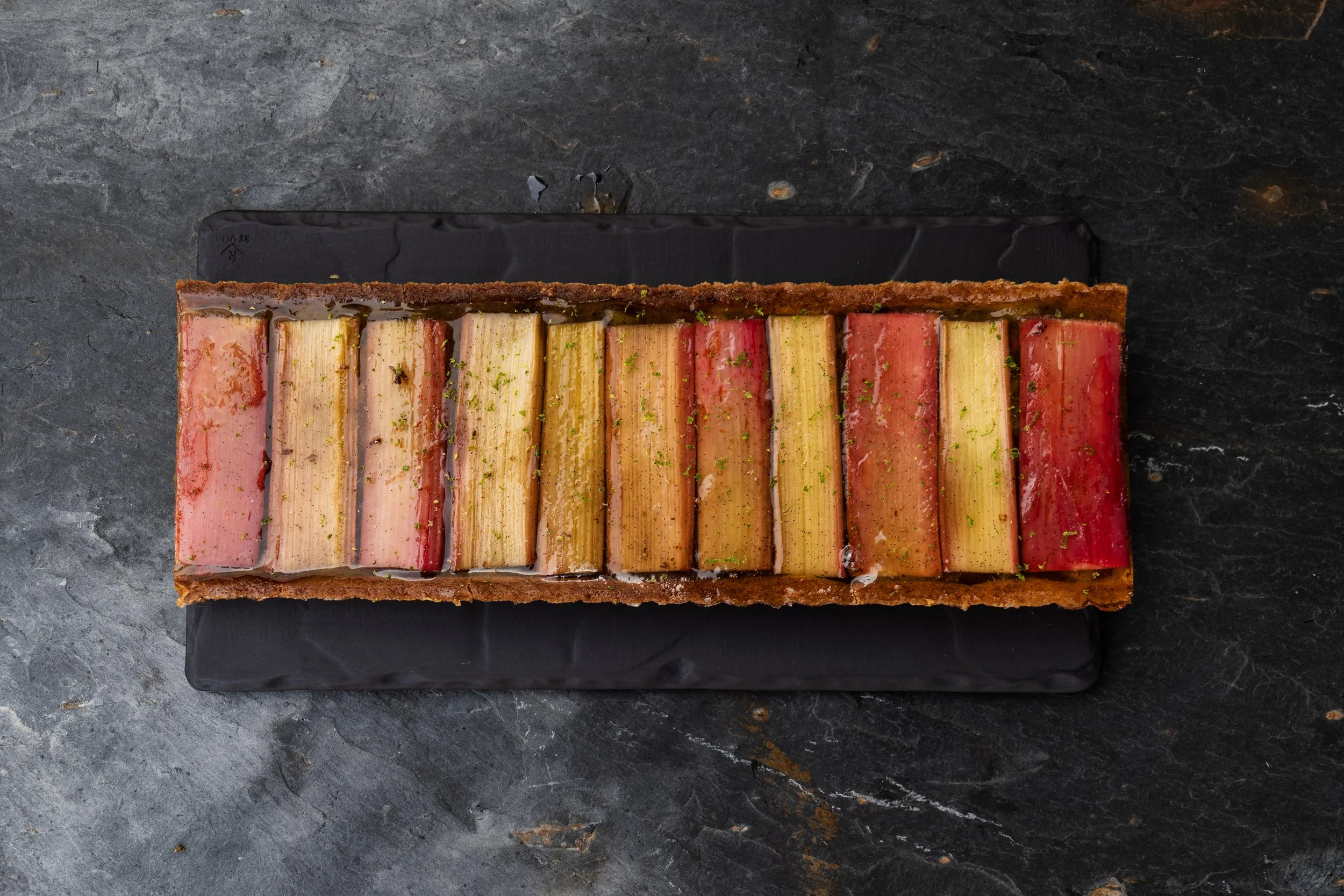 A rectangular multi-layered dessert with colorful layers and a glossy caramel top, garnished with green zest, on a black rectangular plate.
