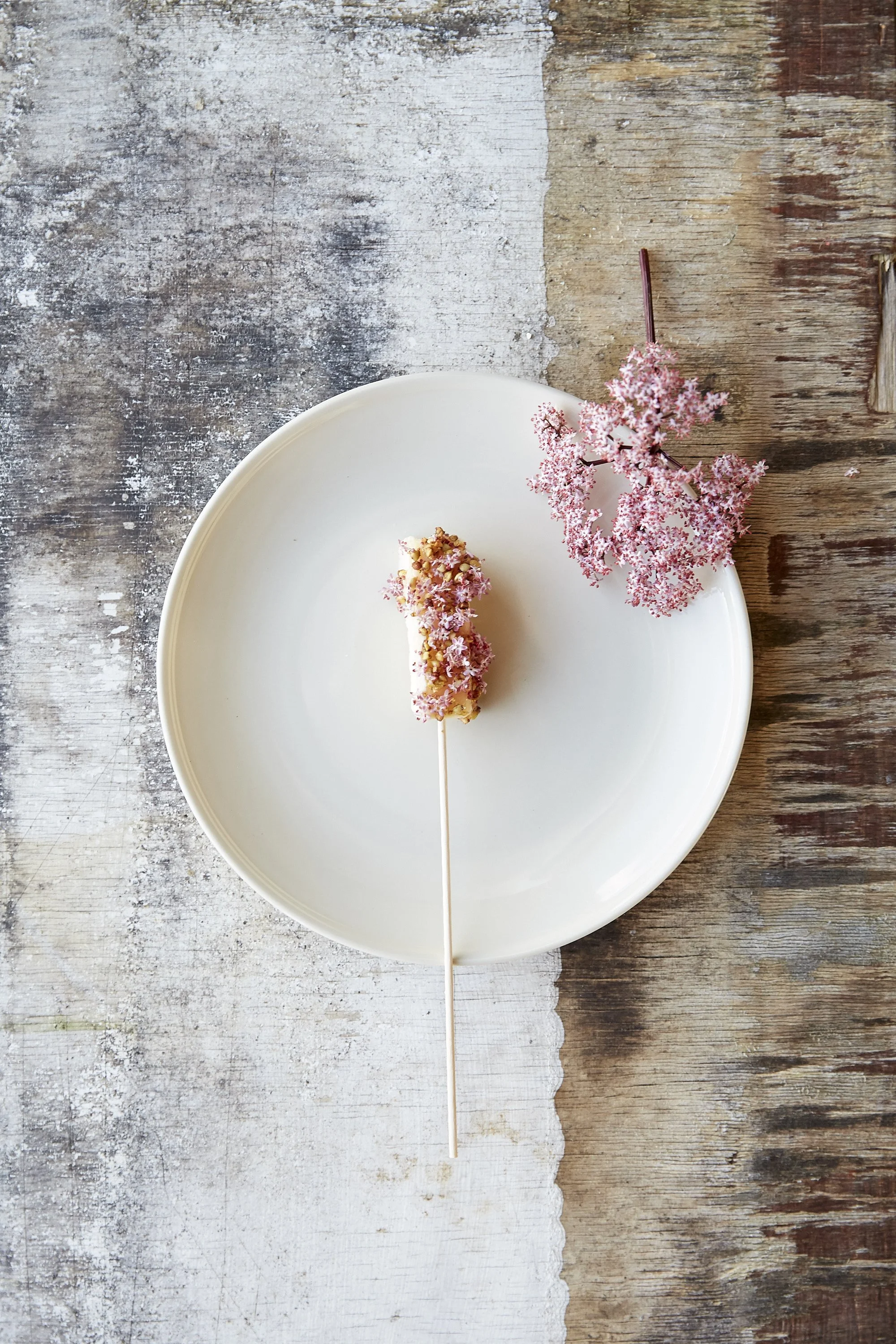 A white plate with a dessert topped with crushed nuts and pink edible flowers, placed on a rustic wooden table with a partially painted surface and a pink flower branch beside the plate.