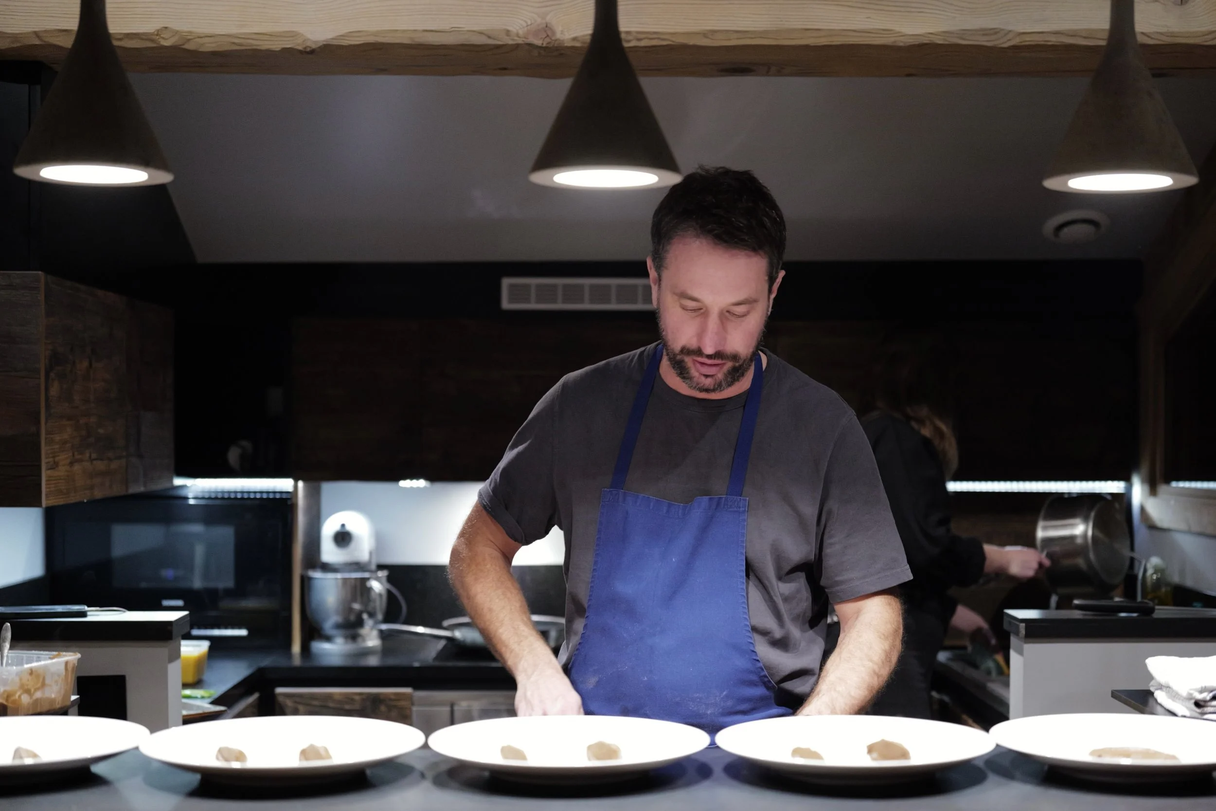 A man wearing a dark t-shirt and a blue apron stands in a modern kitchen with black countertops, preparing food with small white plates in front of him. Overhead, three cone-shaped pendant lights hang from a wooden beam.