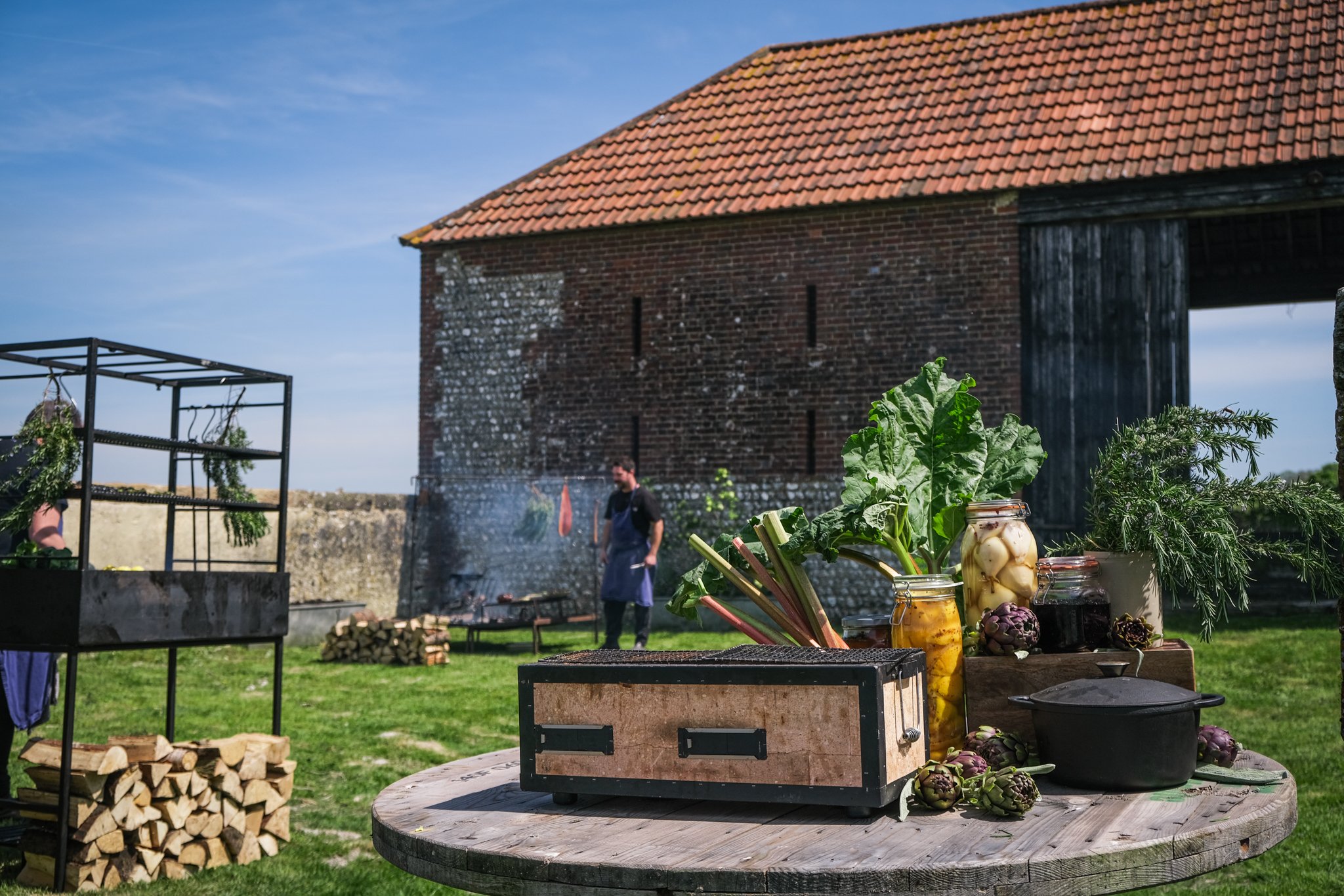 An outdoor market scene with vegetables and jars on a round wooden table, a person grilling in the background, and an old barn with a tiled roof under a blue sky.