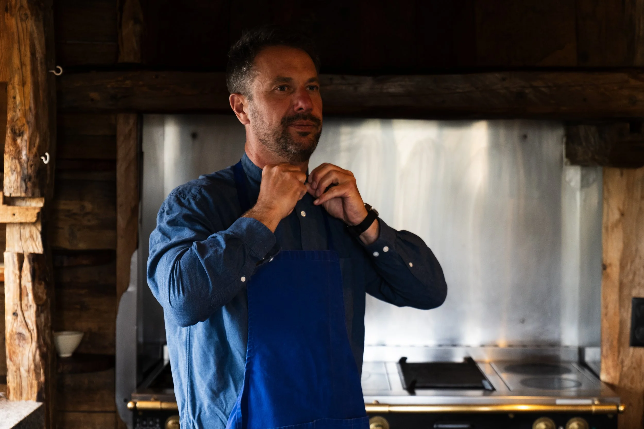 A man adjusting his collar while wearing a blue shirt and apron inside a rustic kitchen with wooden beams and a metallic stove.