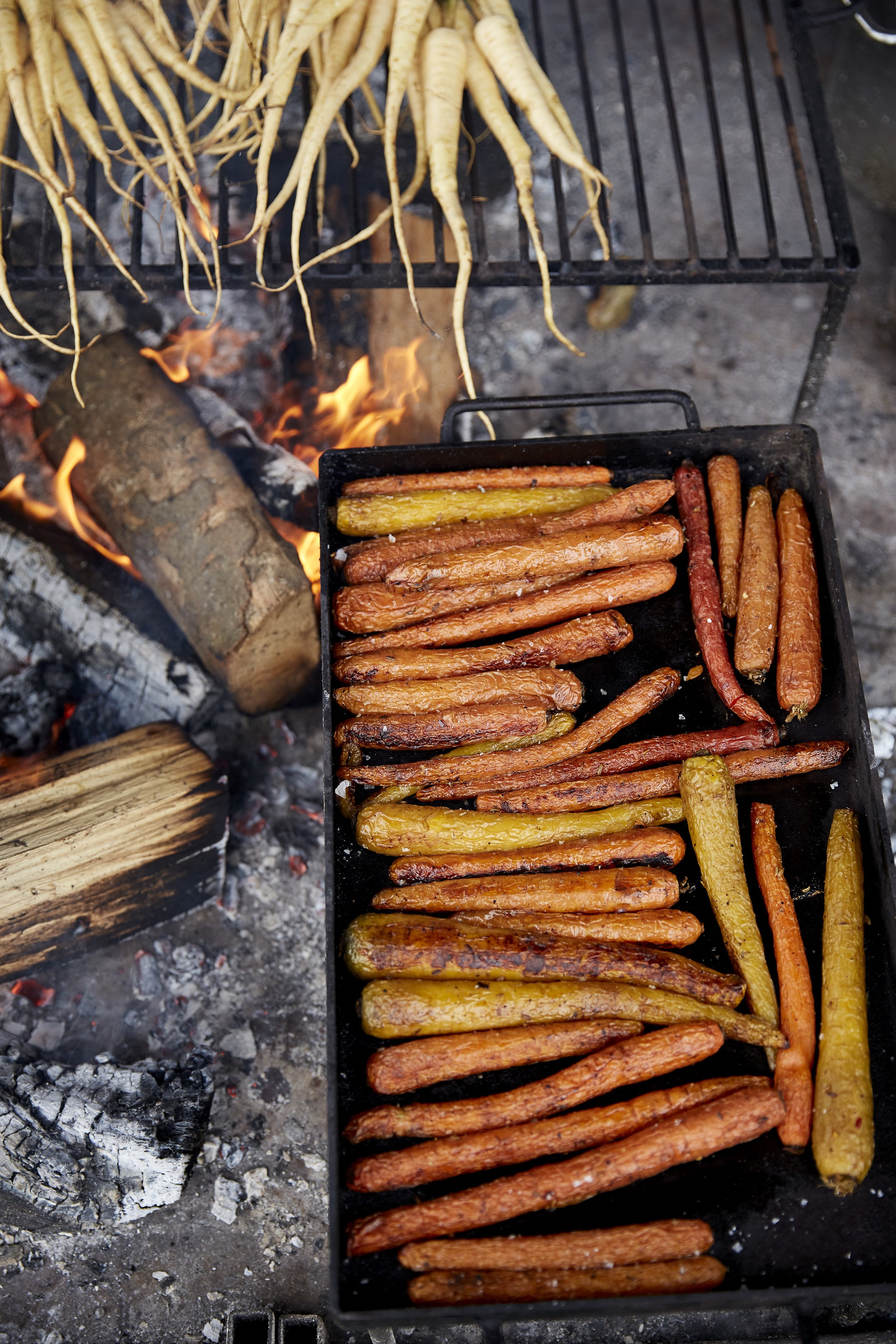 Colorful vegetables, including carrots and yellow squash, grilling on a barbecue over an open flame, with a rack of fresh, raw vegetables nearby.