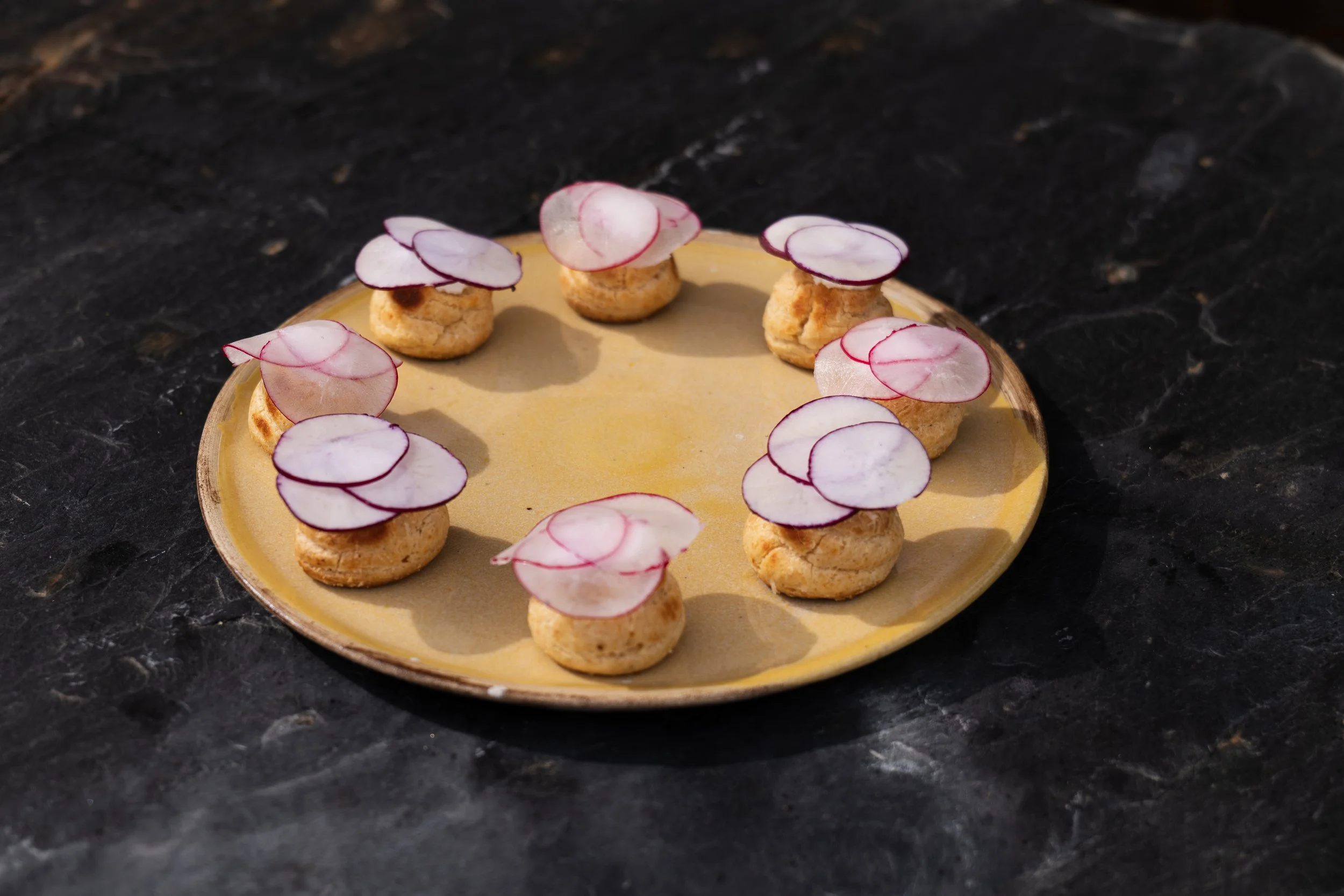 Small pastries topped with thinly sliced radishes arranged on a yellow plate, on a dark stone surface.
