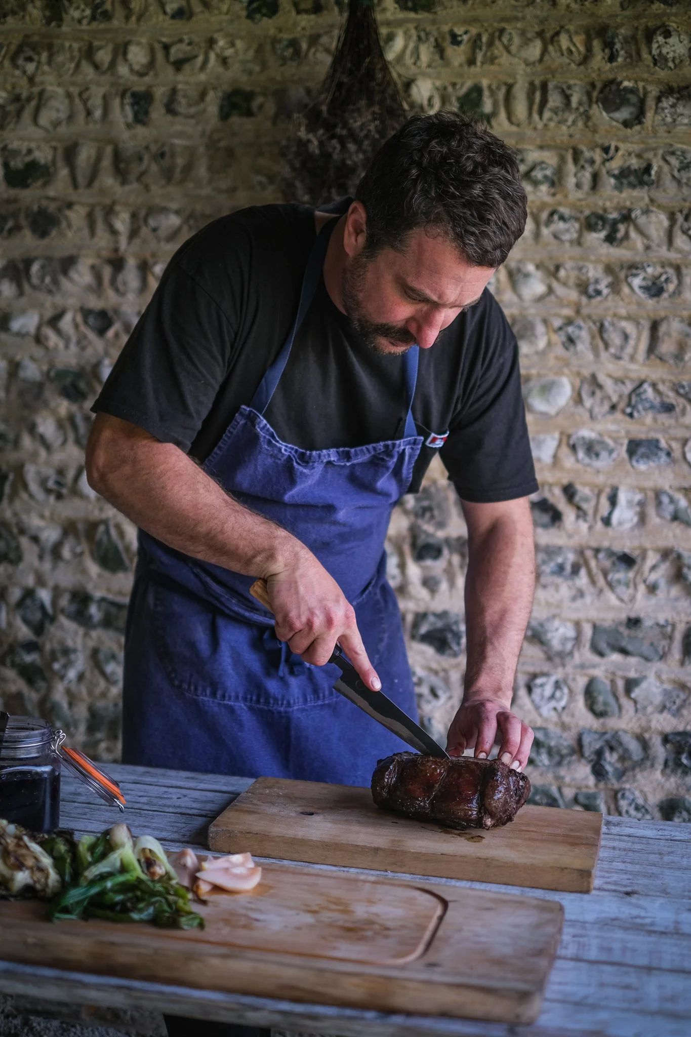 A man wearing a black t-shirt and blue apron is slicing a roasted piece of meat on a wooden cutting board, with a stone wall in the background.