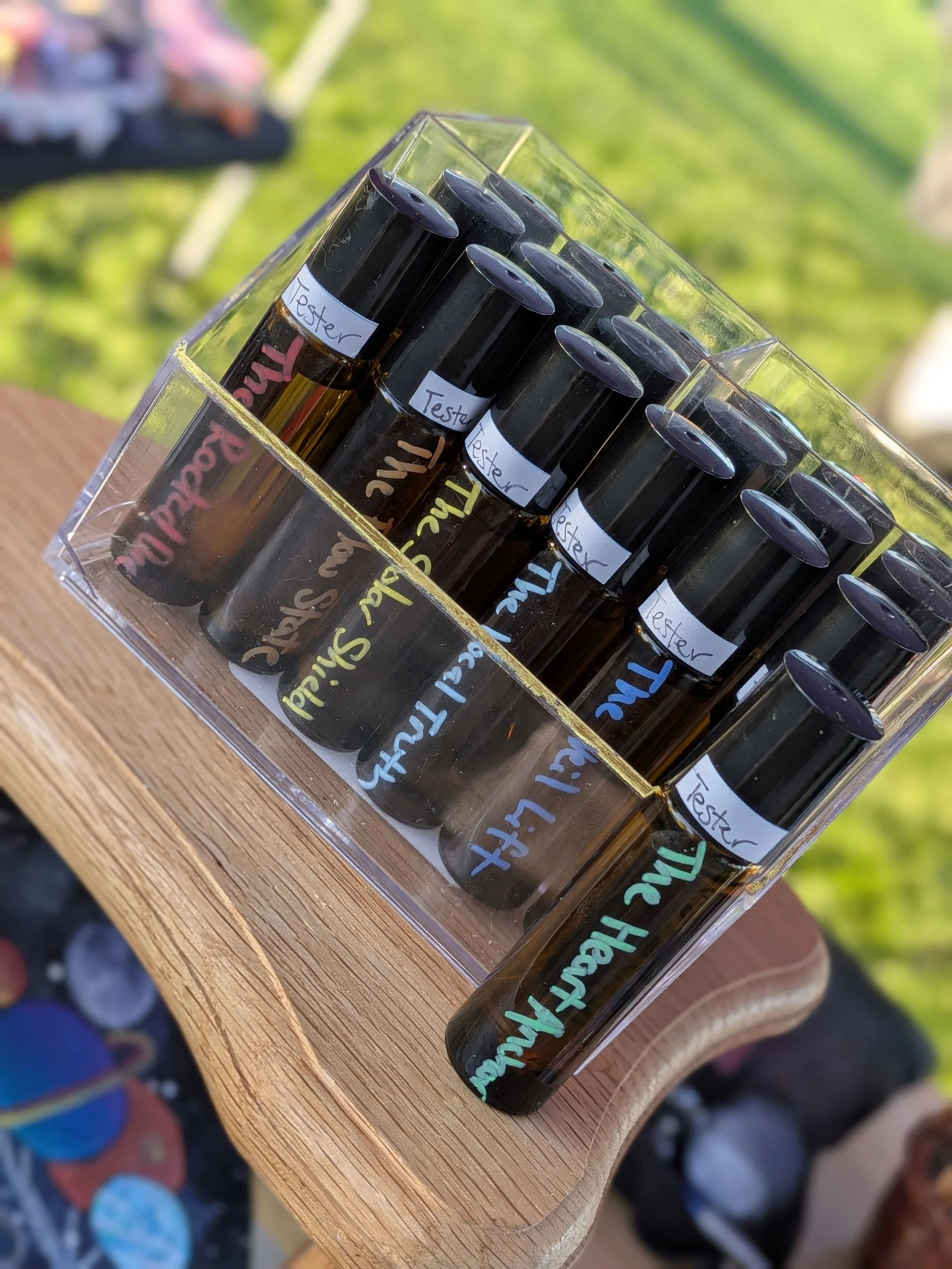Multiple test tubes with black caps in a clear plastic container, each labeled with a handwritten 'Tester' label in various colors, placed on a wooden surface outdoors with greenery in the background.