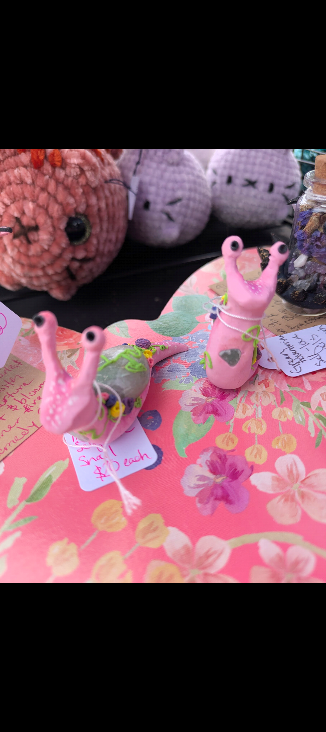 Colorful handcrafted snail figurines with black eyes, placed on a pink floral tablecloth at a craft market.