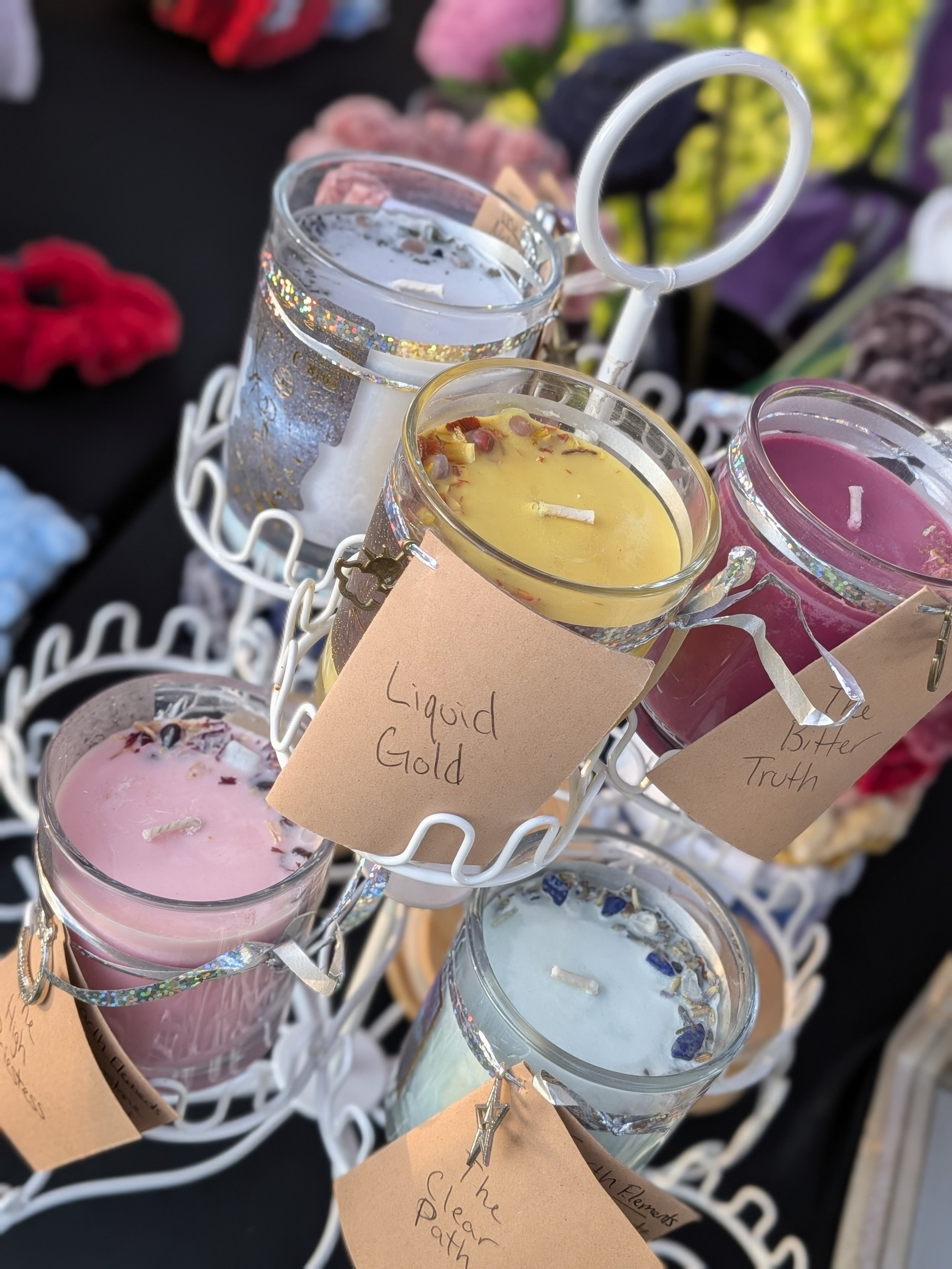Assorted candles in small glass containers with handwritten labels, arranged on a white decorative stand, with blurred background of various textiles and flowers.
