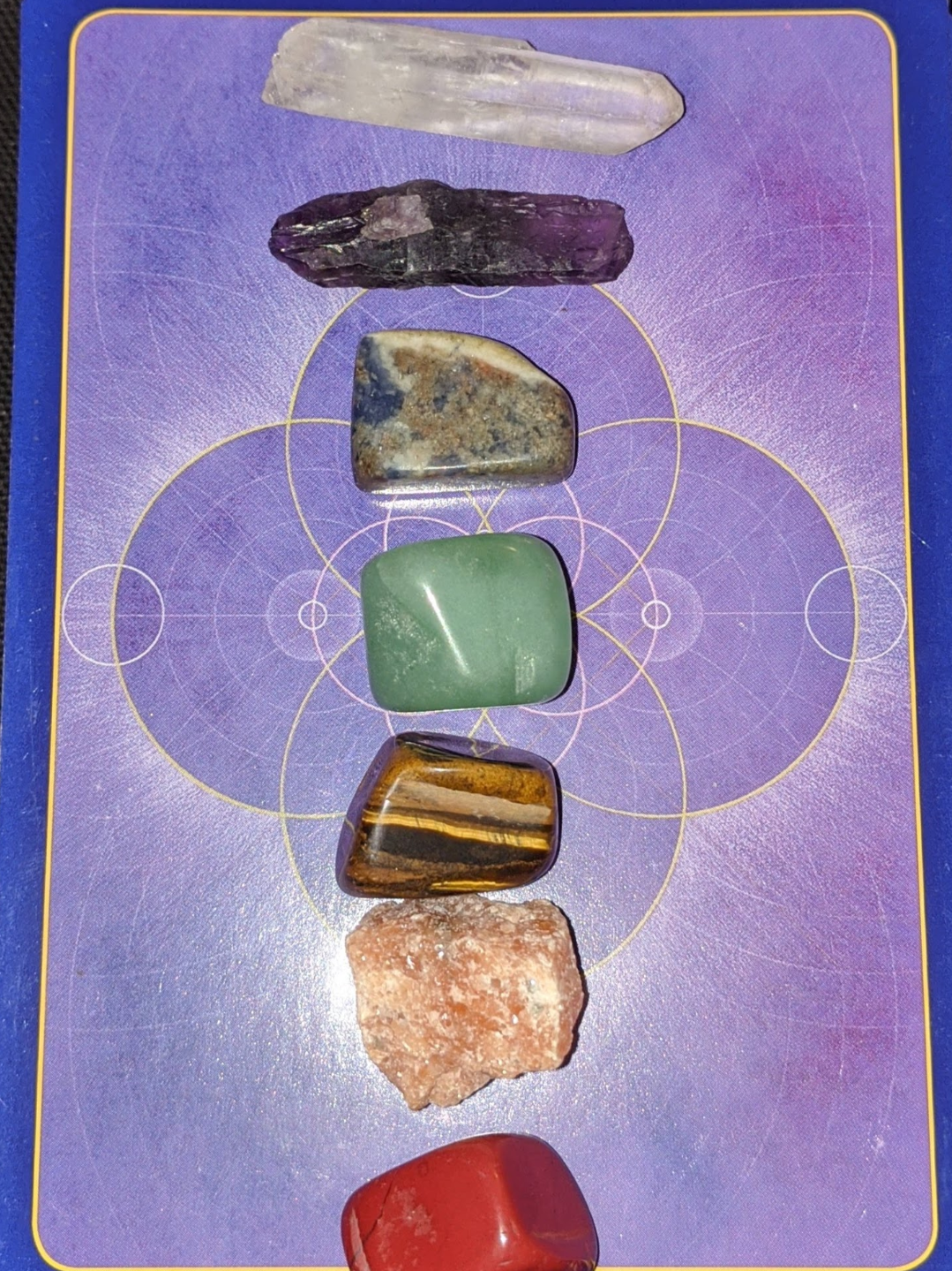 A collection of various polished and raw gemstones aligned vertically on a purple tarot card background with gold geometric patterns.