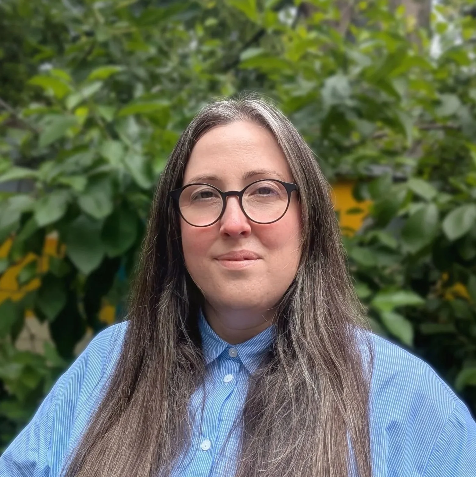 Portrait of a woman with long brown hair, glasses, and a blue striped shirt, standing outdoors with green foliage in the background.