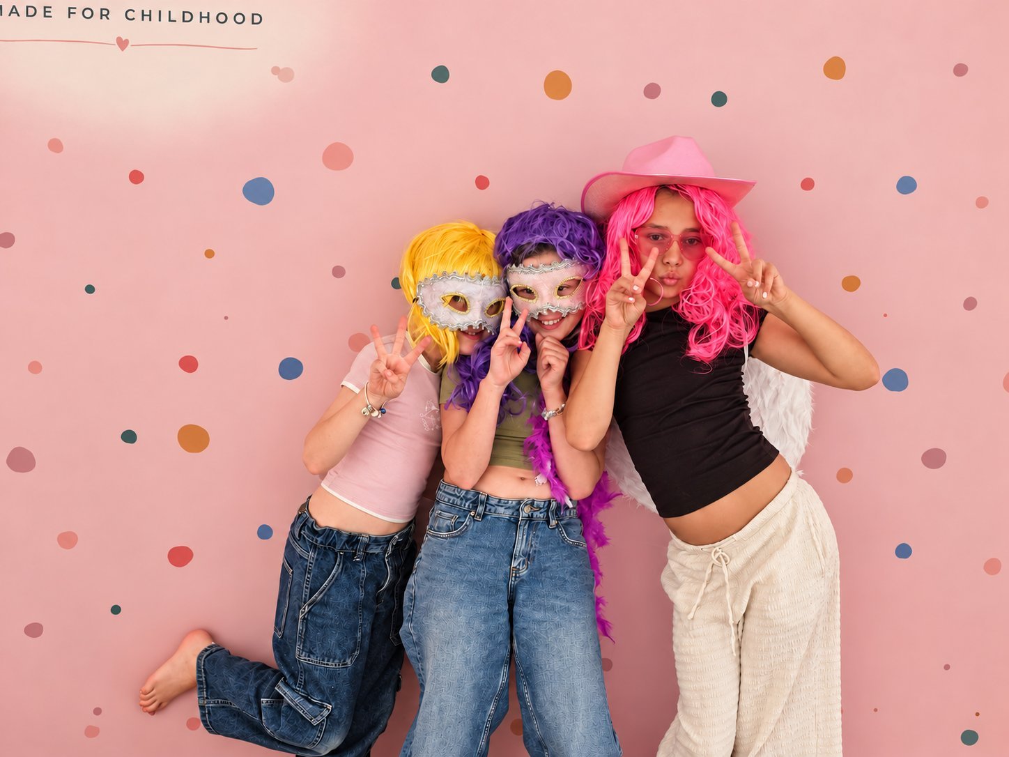 Three girls in colourful wigs and masquerade masks posing at the Willow & Mae photo wall
