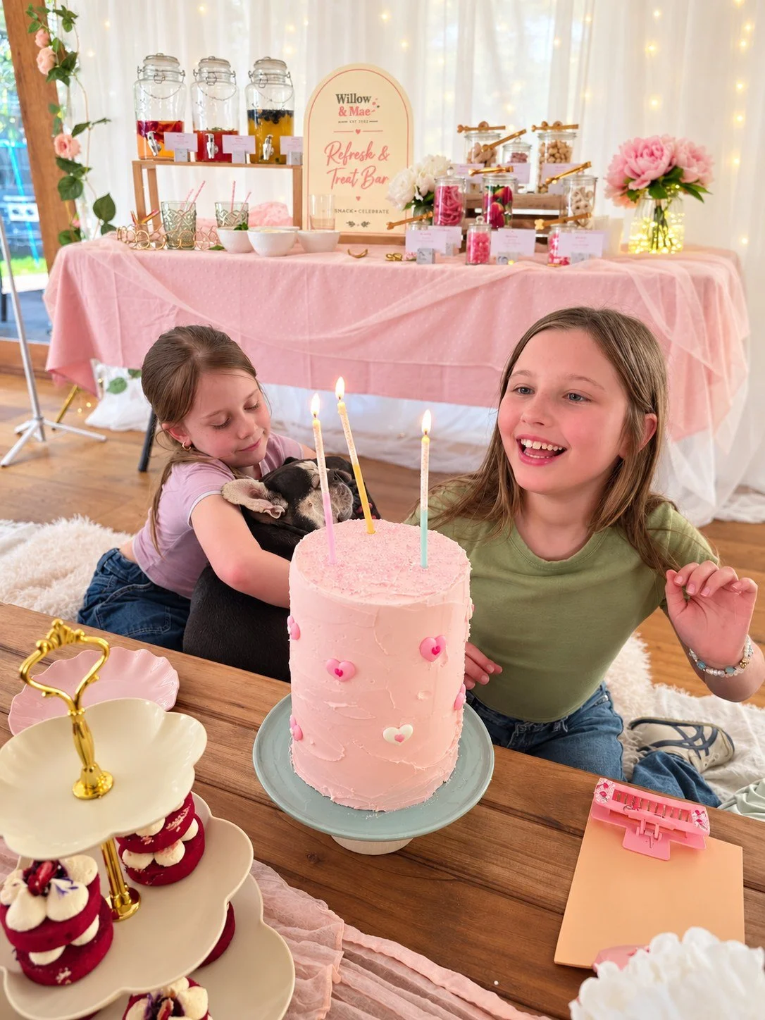 Two girls laughing beside a pink birthday cake with candles and red velvet cupcakes