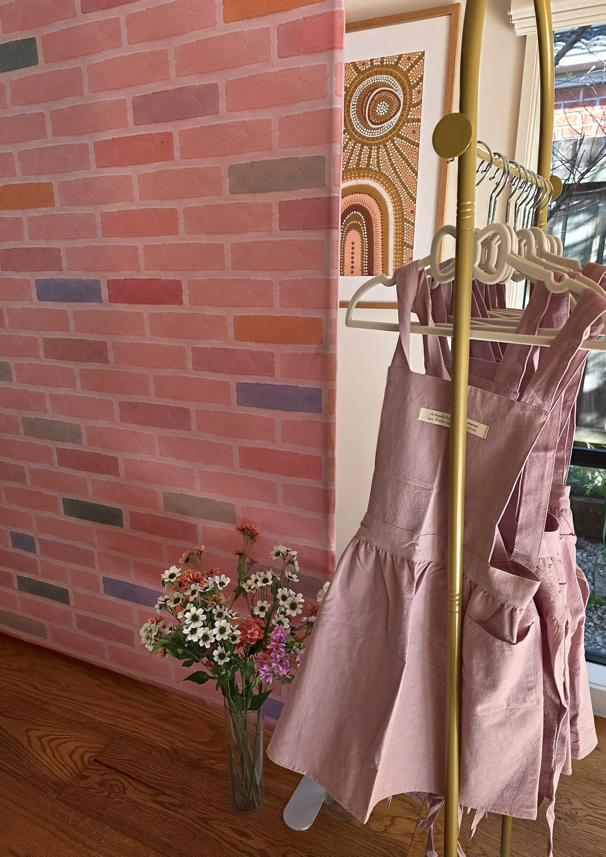 Lilac craft apron waiting for a guest on a gold rack beside a pink brick wall and fresh flowers