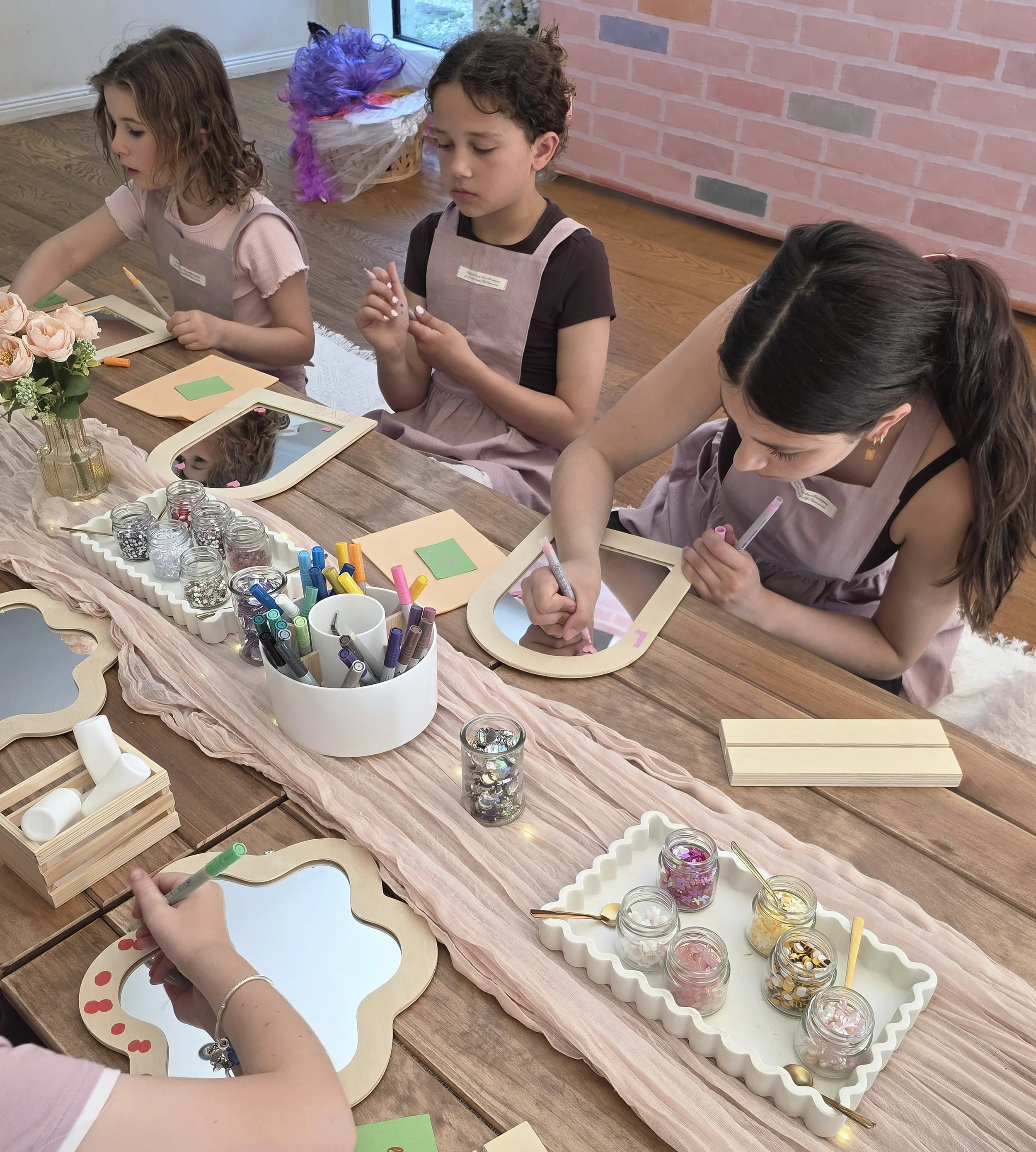Girls in aprons decorating wooden mirrors with paint pens at a Creative Studio craft party
