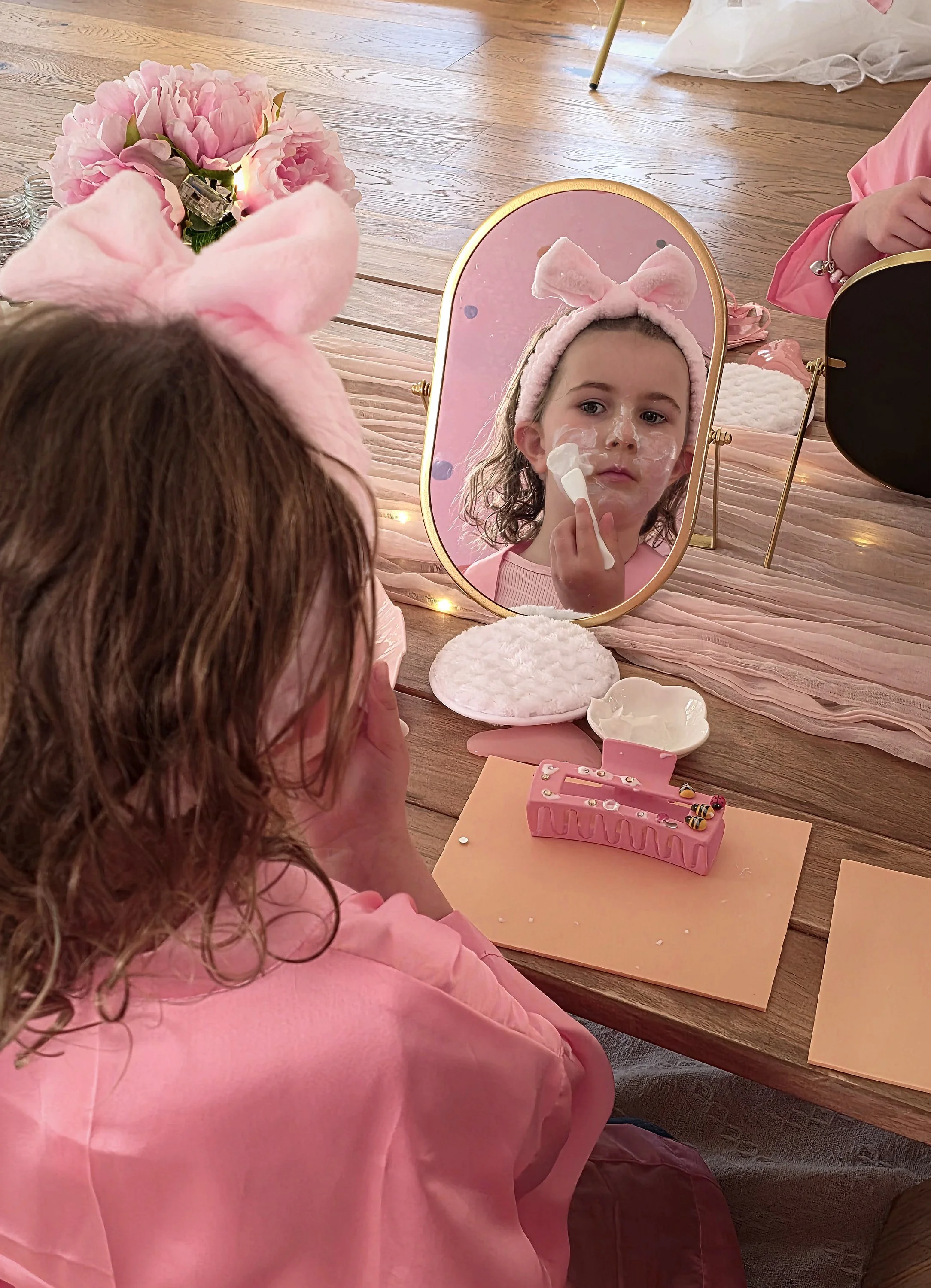 Spa party guest in a bow headband applying face mask in a gold mirror