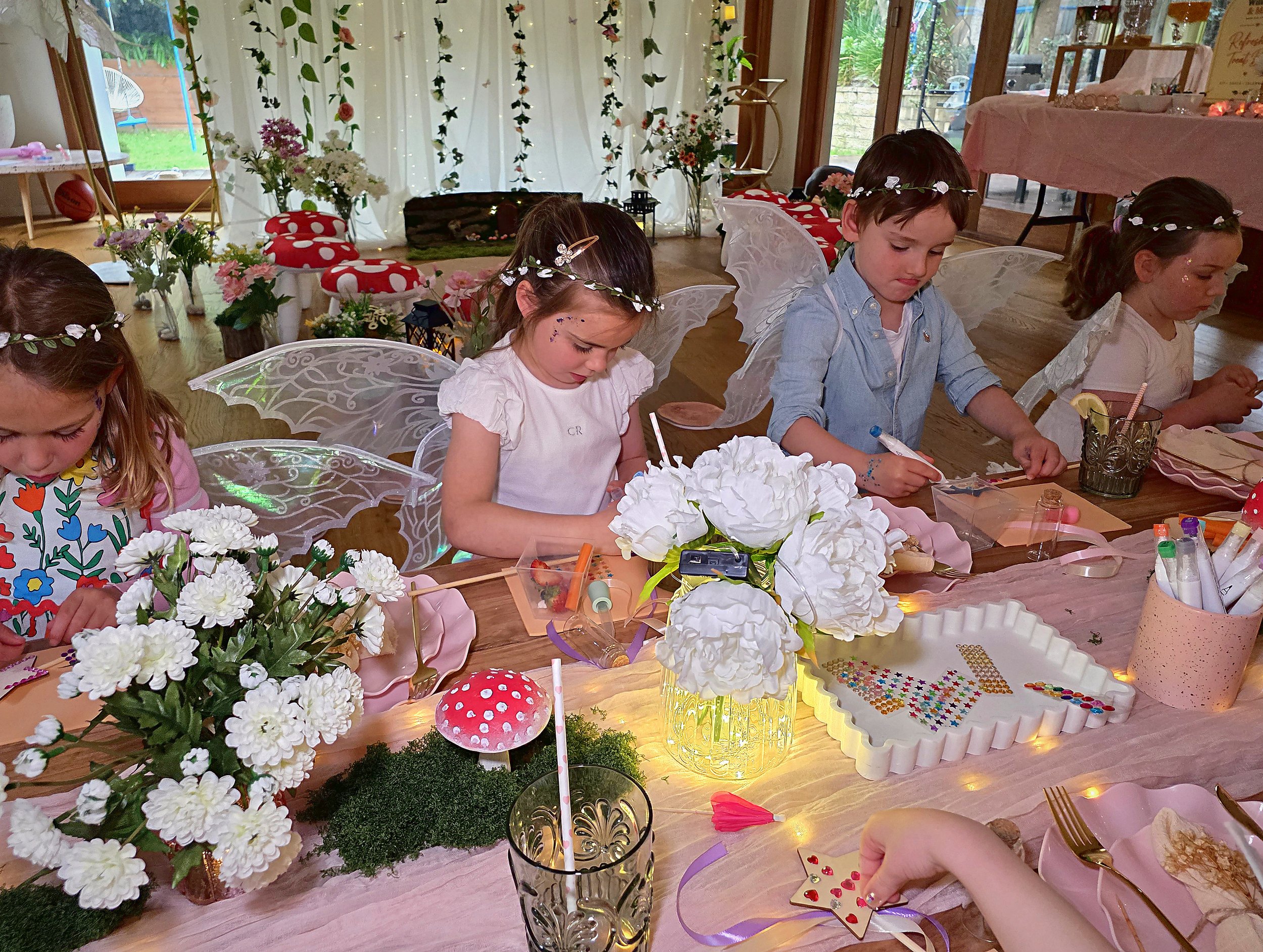 Fairy party guests decorating wands at a table with white flowers, toadstools and gem trays