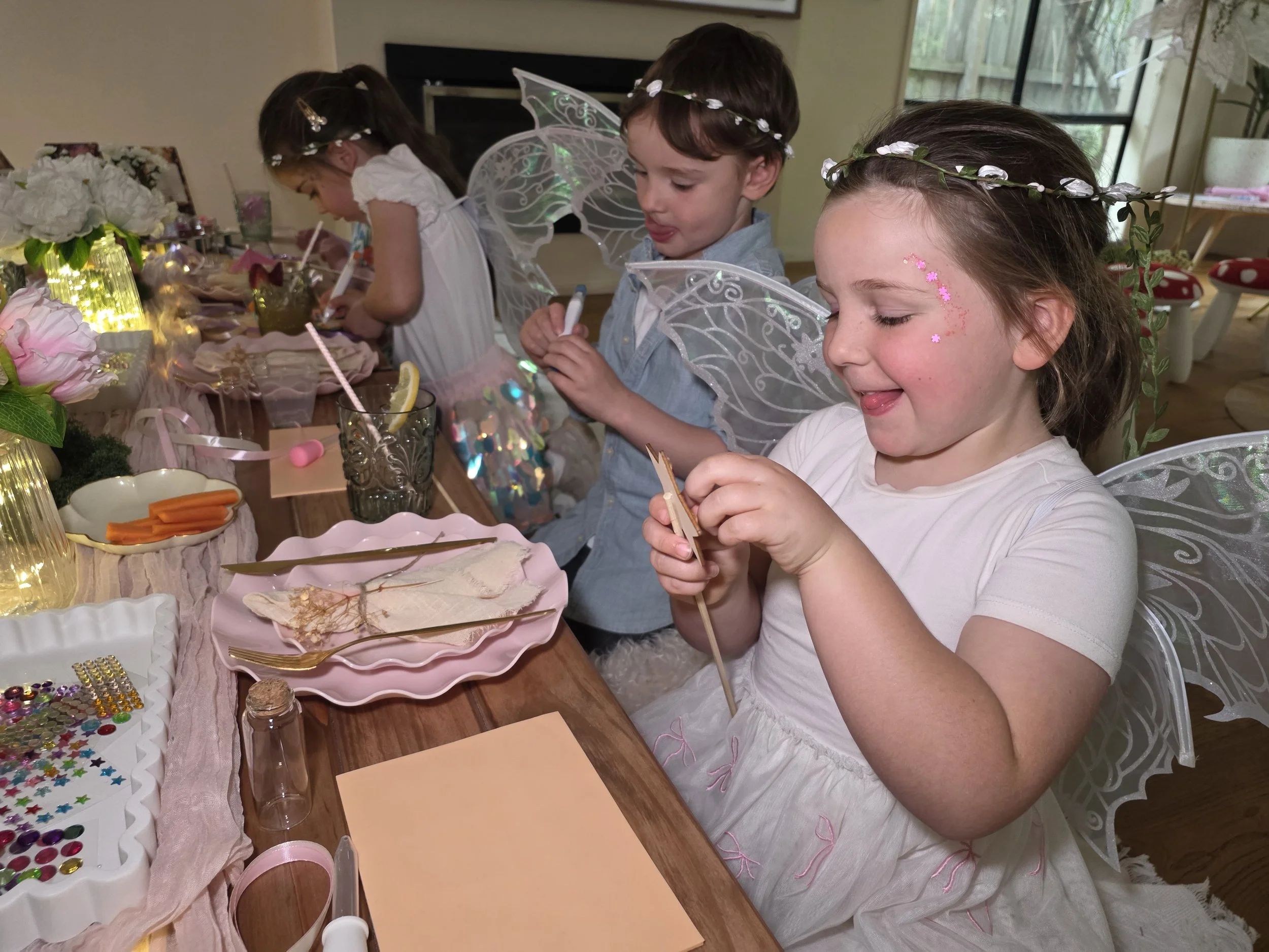Smiling children in fairy wings and flower crowns making magic wands at the party table