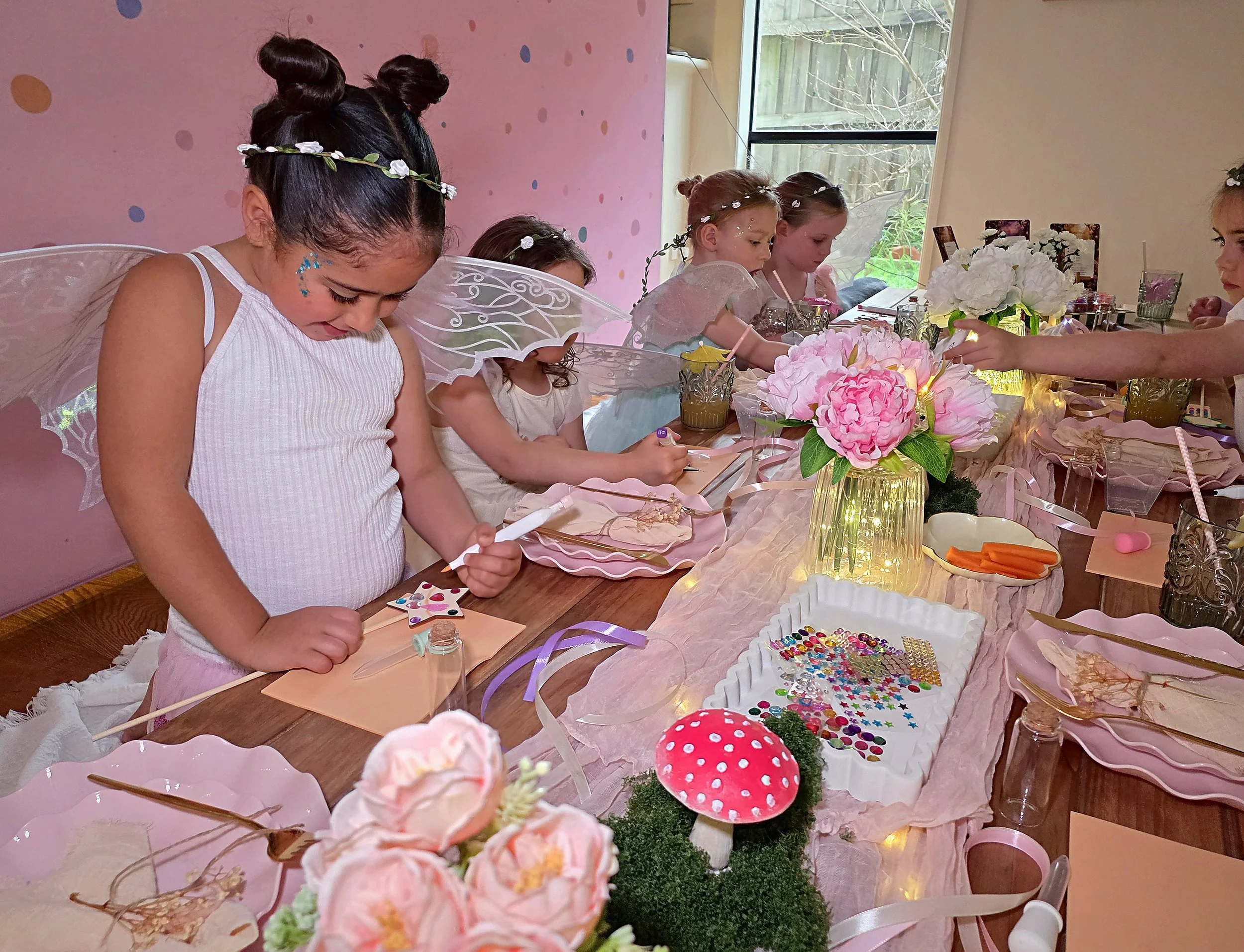 Girl with face glitter and fairy wings decorating her wand beside pink peonies