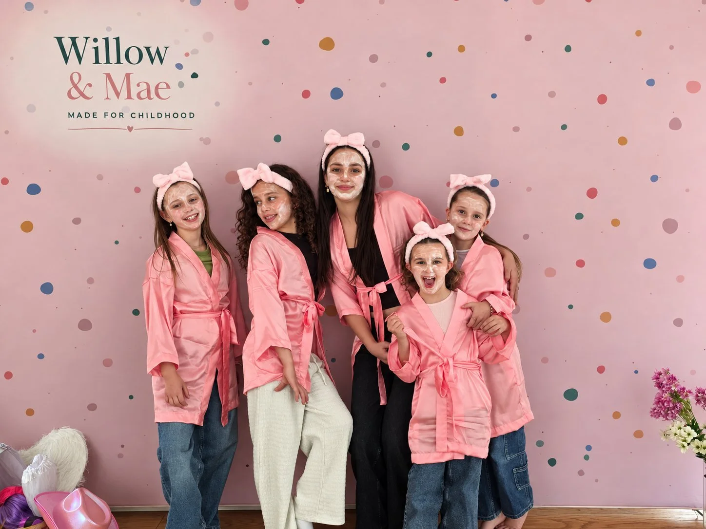 Five girls in pink spa robes, bow headbands and face masks posing at the Willow & Mae backdrop
