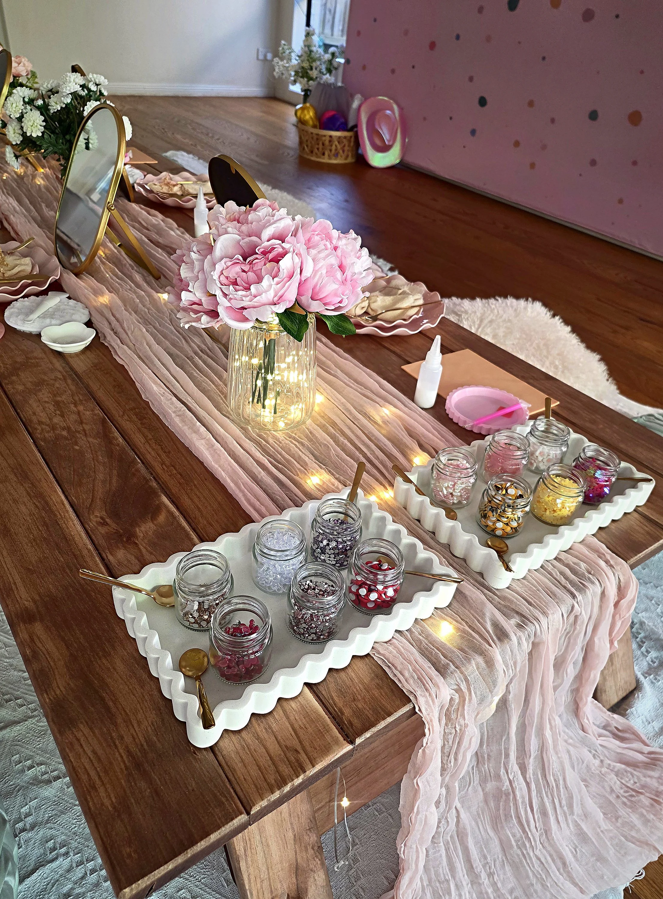 Gem and embellishment trays, pink peonies and fairy lights set out on a spa party table