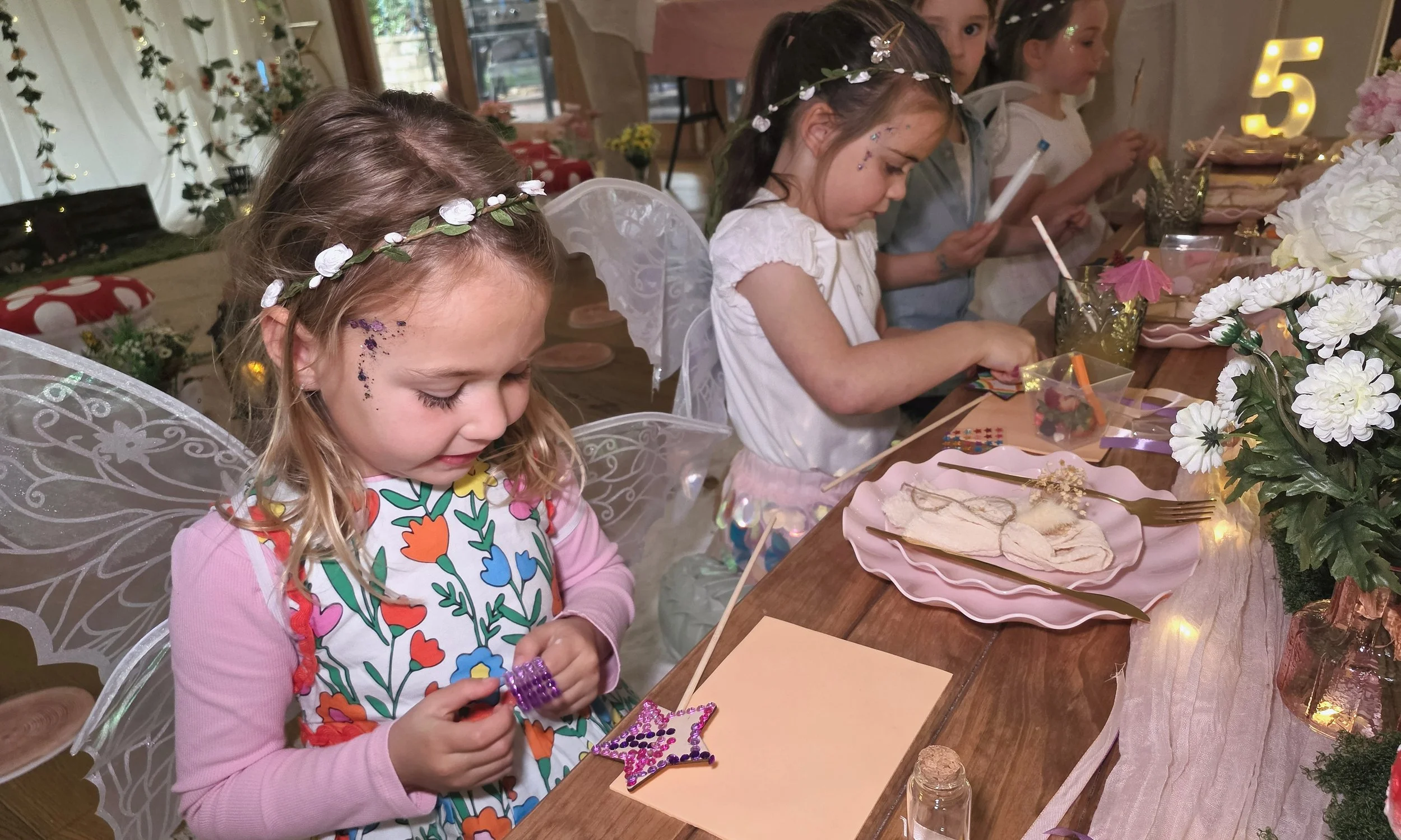 Little fairy with face gems and a flower crown decorating her star wand at the party table