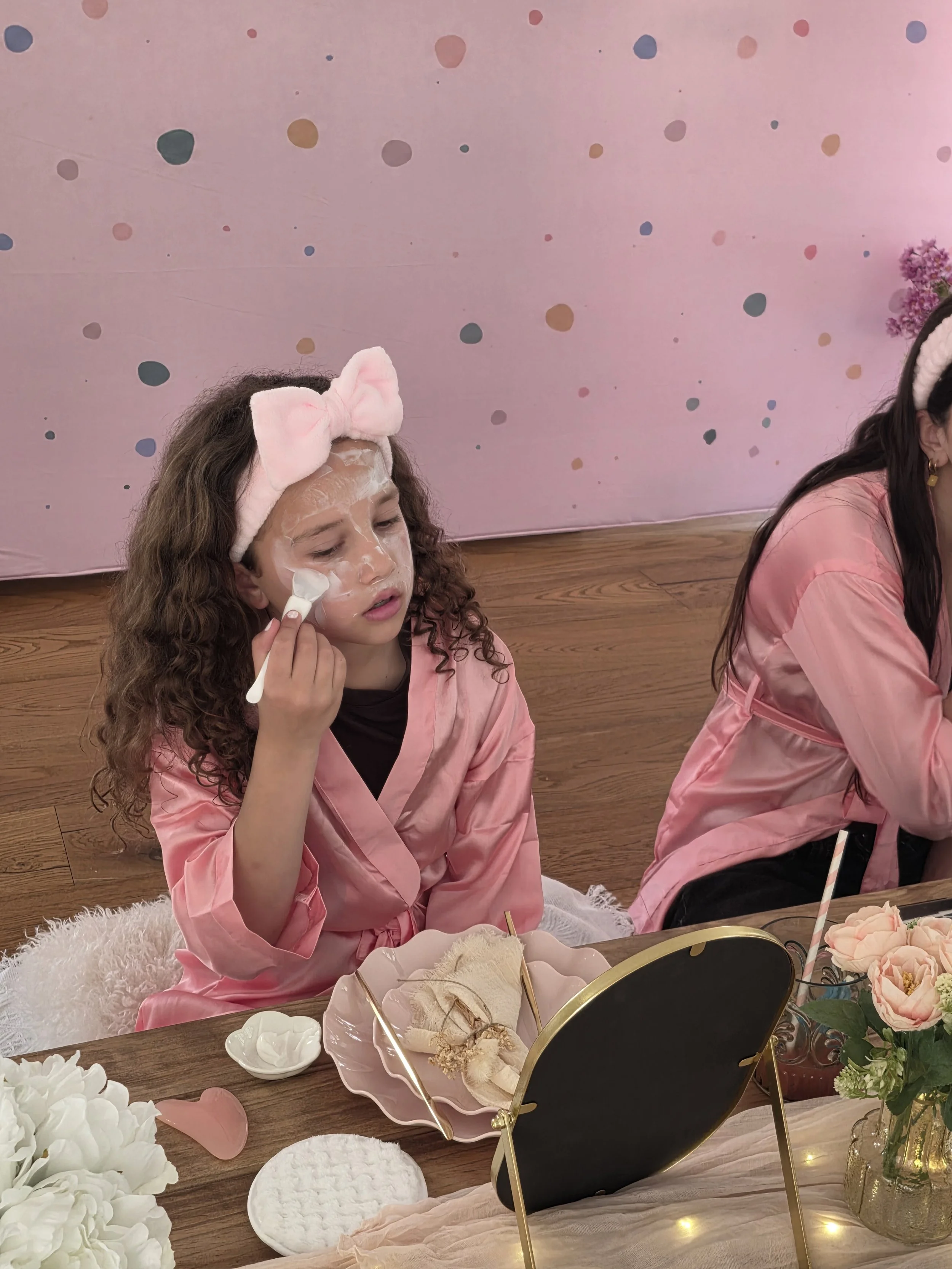Girl in a pink robe and bow headband brushing on a face mask at a spa party