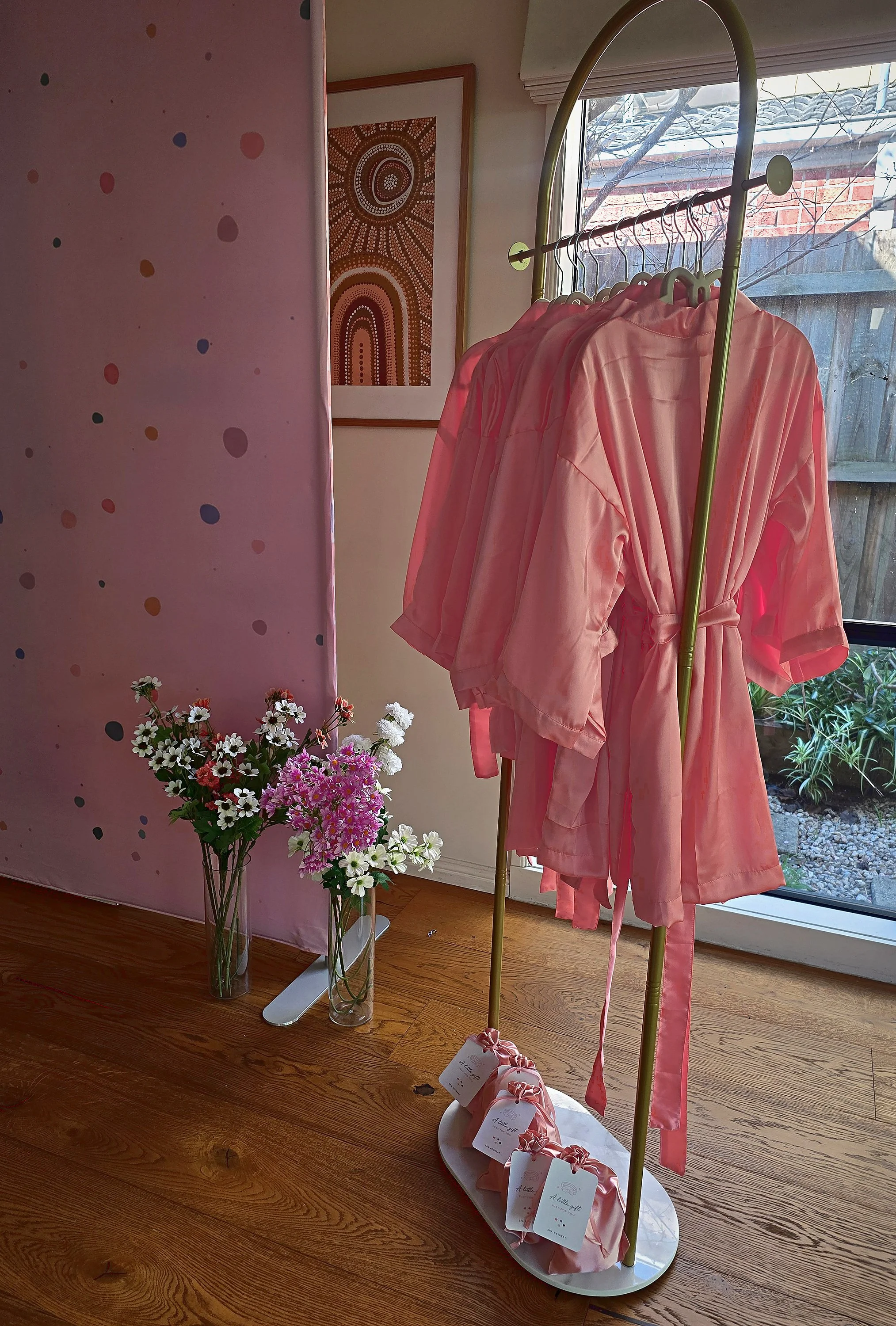 Pink satin spa robes hanging on a gold rack beside flowers and gift bags