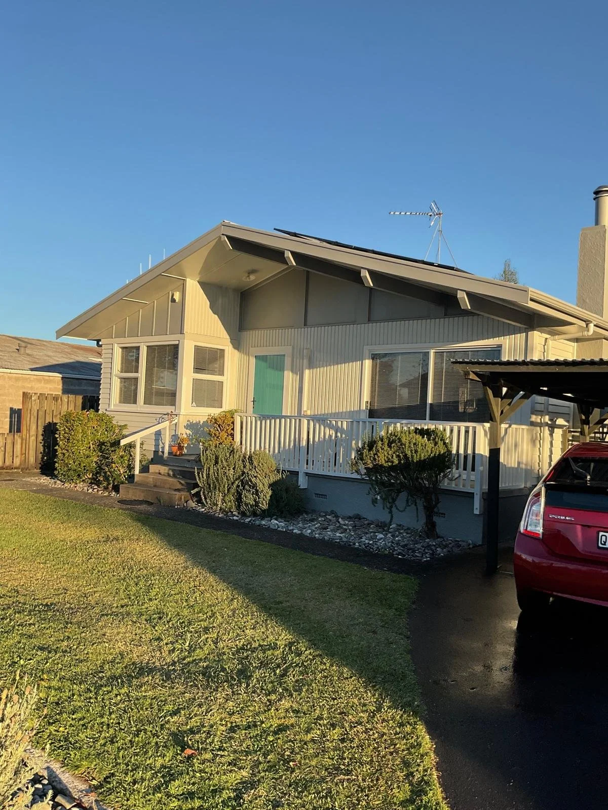 A suburban house with a front porch, light yellow siding, a small staircase, and a manicured lawn. There is a red car parked under a black carport on the driveway. The house has a pitched roof with an antenna, and the sky is clear and blue.