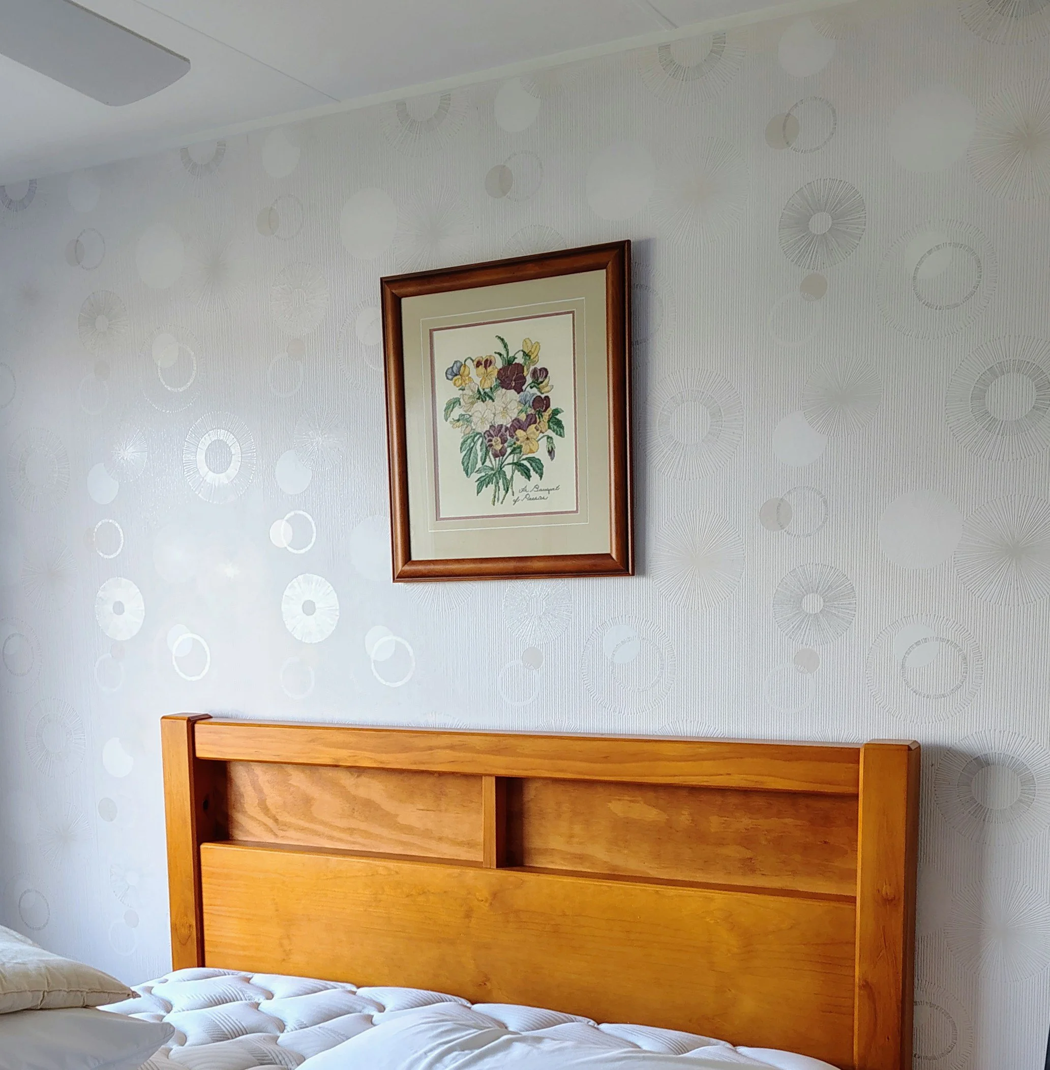 A bedroom with a wooden headboard, a bed with white bedding, and a framed botanical print on the wall. The wall has a patterned wallpaper with circular and radiating line designs.