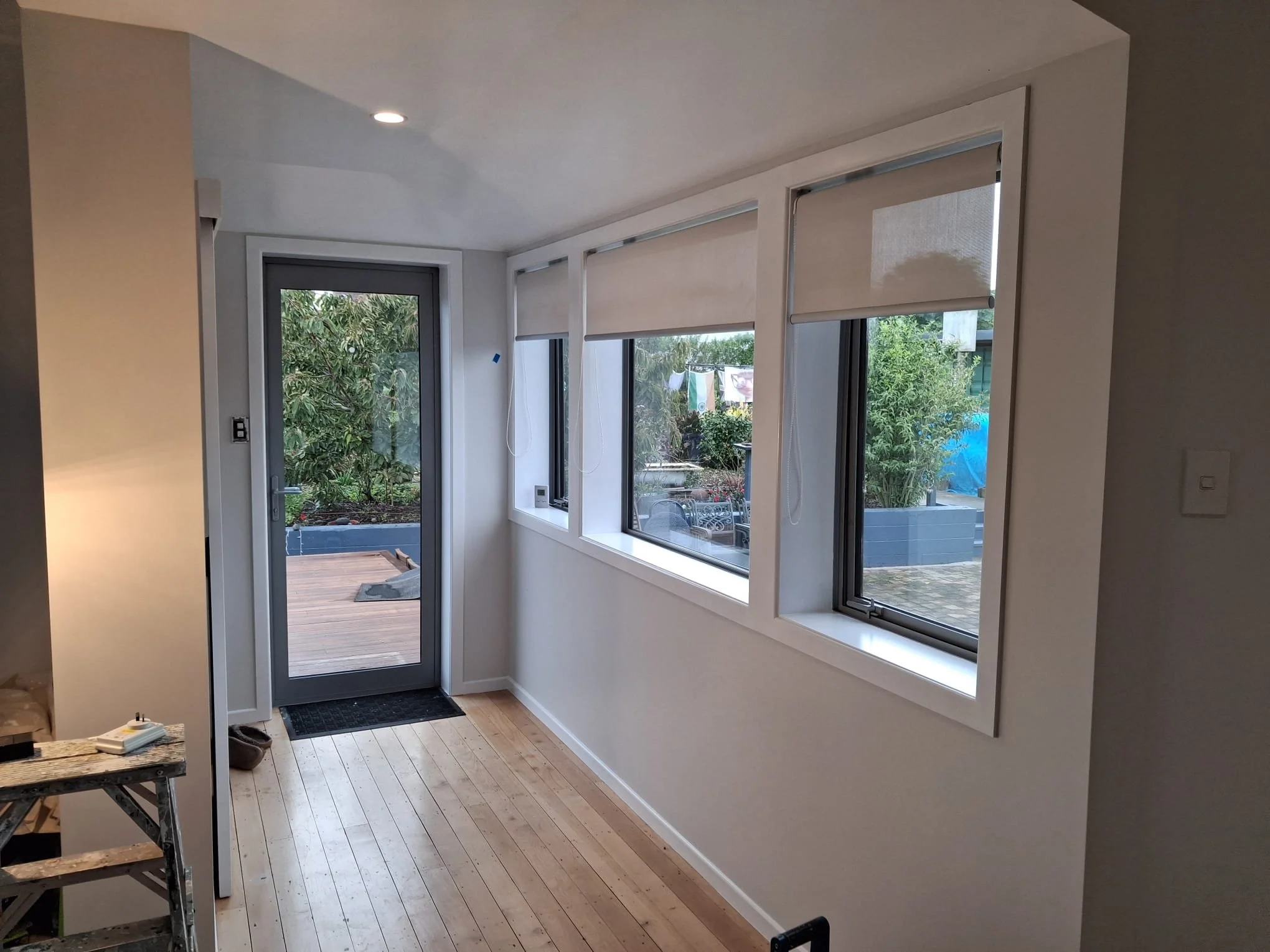 Inside view of a modern home entryway with hardwood flooring, large windows with roller blinds, and a door leading to an outdoor deck with greenery.