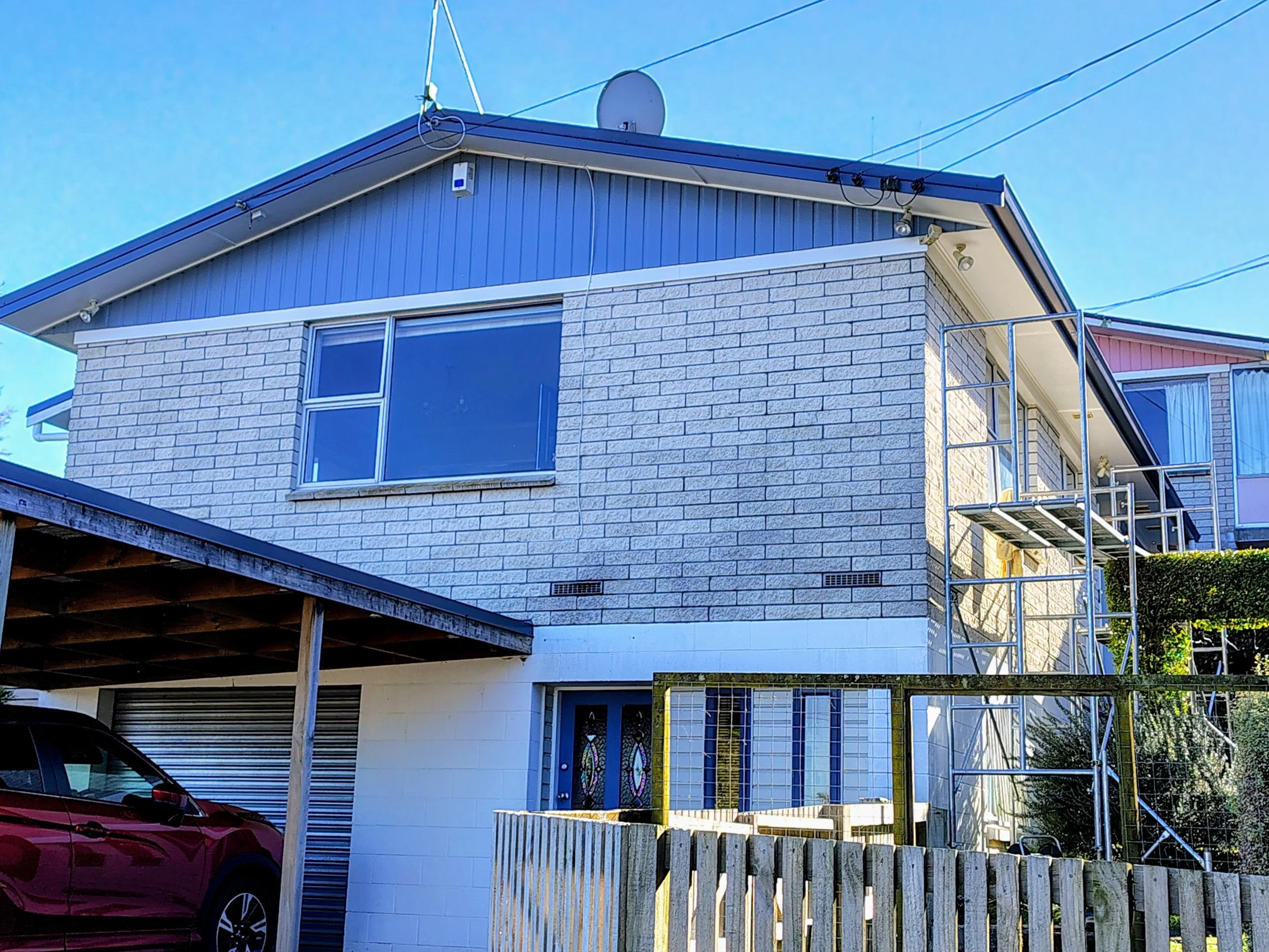 A two-story house with a brick exterior and large windows on the upper floor. There is a small balcony with metal railing on the right side. Scaffolding is set up on the right side of the house, and part of a red car is visible on the lower left corner. The sky is clear and blue.