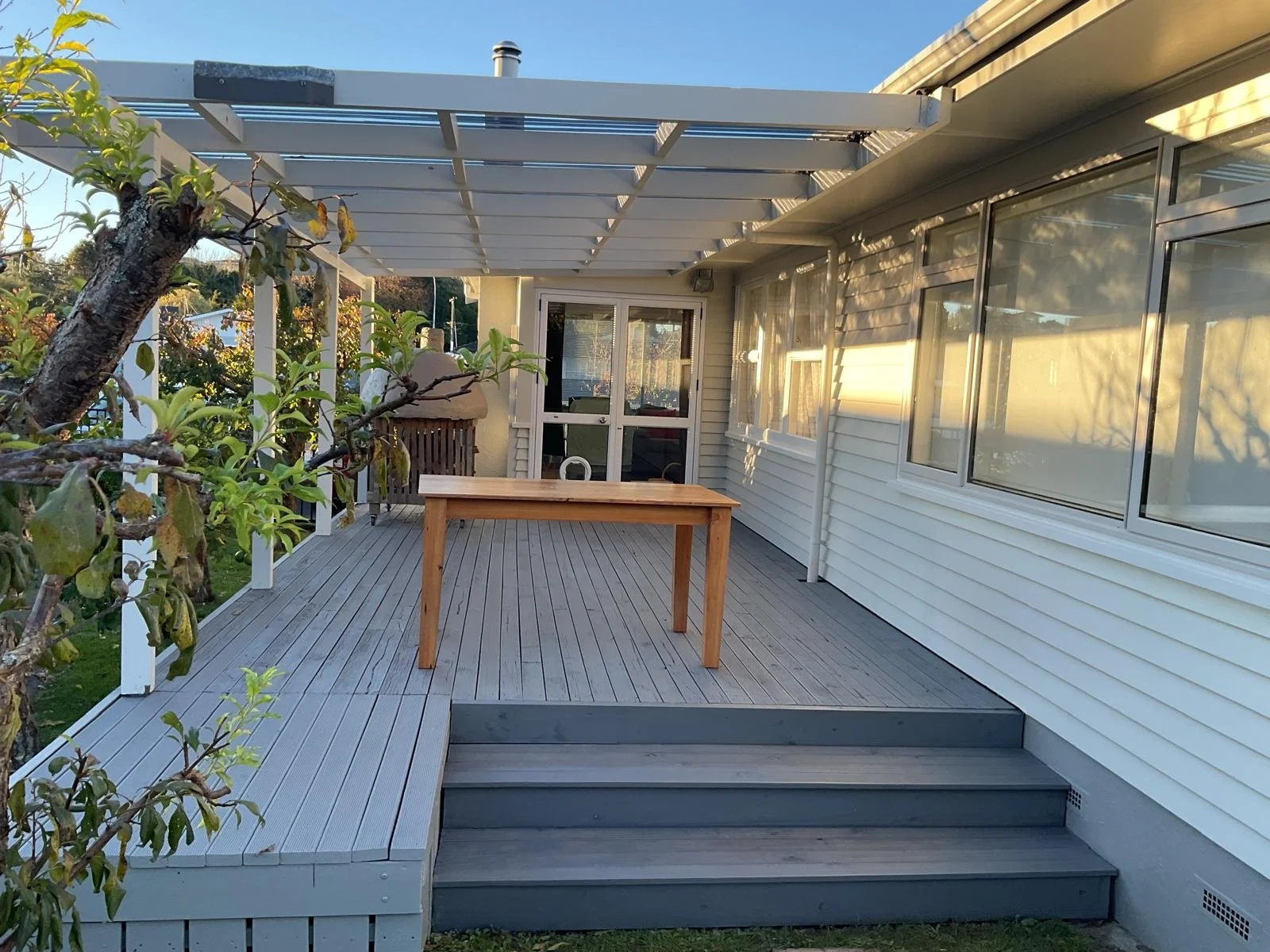 Photo of a backyard porch with gray wooden decking, a small wooden table, white siding house, large windows, tree with green leaves, and a clear sky.
