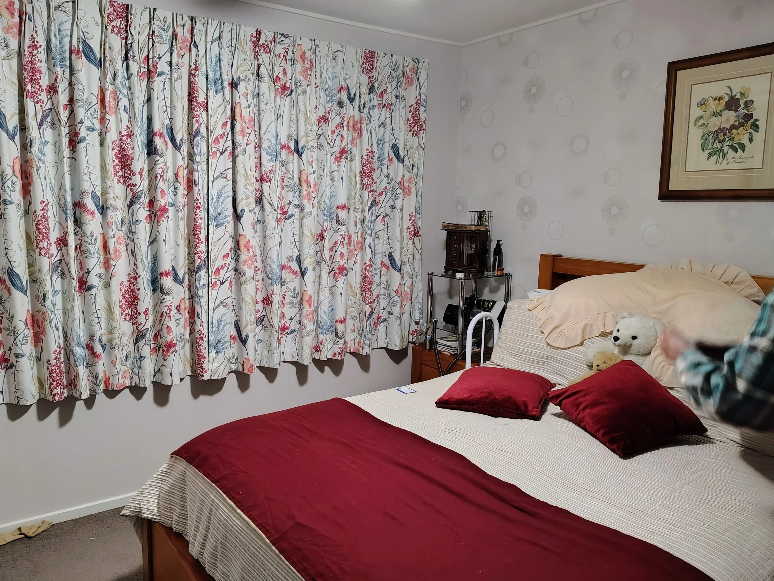 A bedroom with two beds covered with red duvets, one bed has a stuffed white bear and pillows. There are floral patterned curtains, a framed flower picture on the wall, and small furniture including a nightstand with books and a soap dispenser.