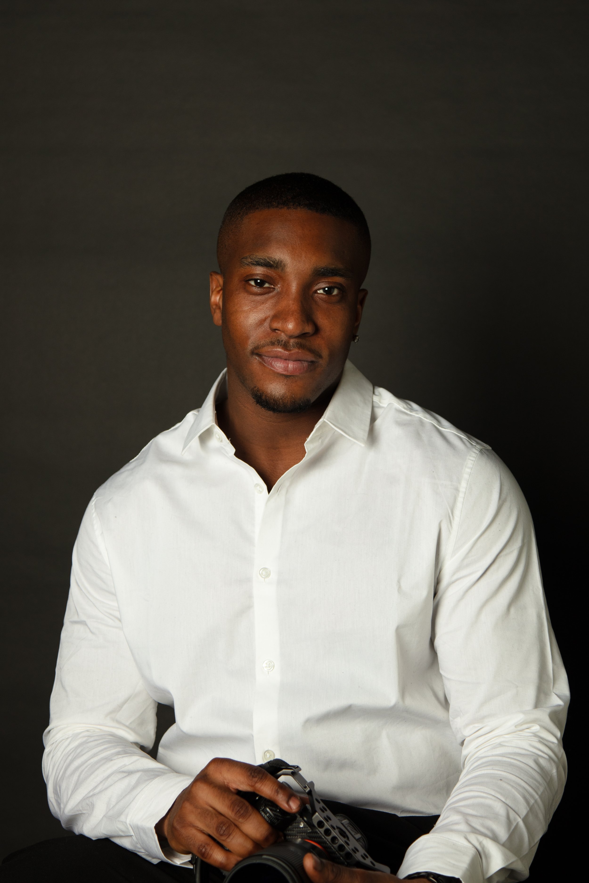 A young man with short hair and a mustache, wearing a white dress shirt, sitting against a dark background, holding a camera in his hands.