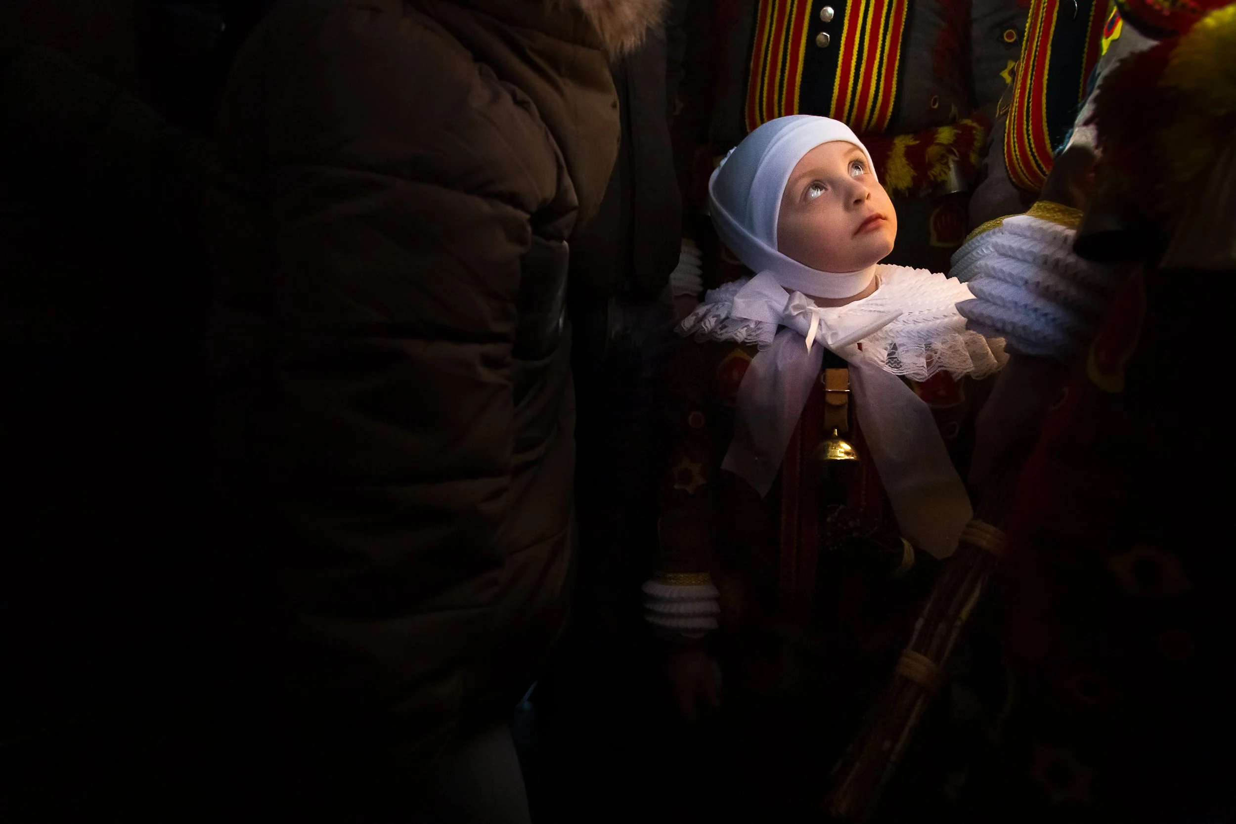Un jeune enfant regarde vers le haut dans une scène sombre, entouré de personnes portant des vêtements traditionnels colorés.
